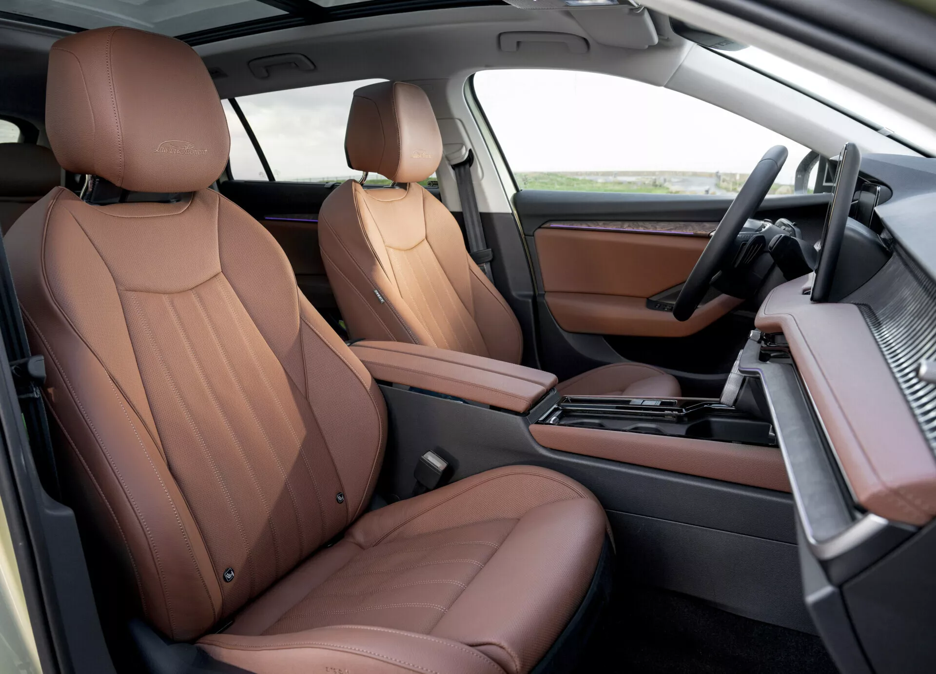 Luxurious brown leather seats and modern dashboard in the 2024 Skoda Superb Combi's front cabin, showcasing automatic gear shift, infotainment system, and wood-finish accents from a top-down passenger perspective