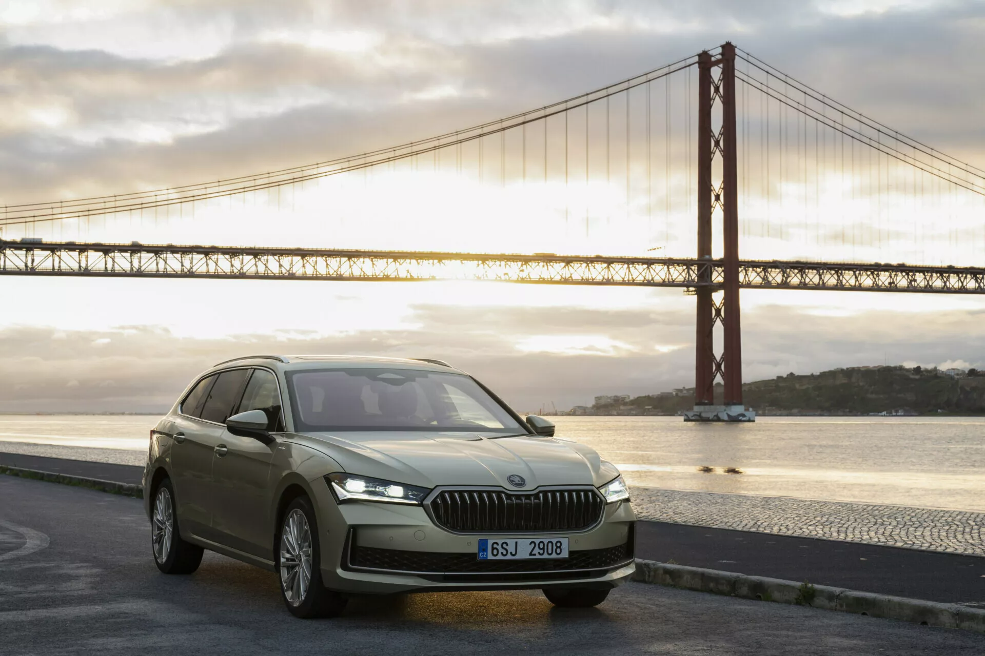 2024 Skoda Superb Combi luxury SUV at dawn, showcasing modern design features like vertical front grille, sleek LED headlights, contoured hood, and aerodynamic profile near iconic bridge.