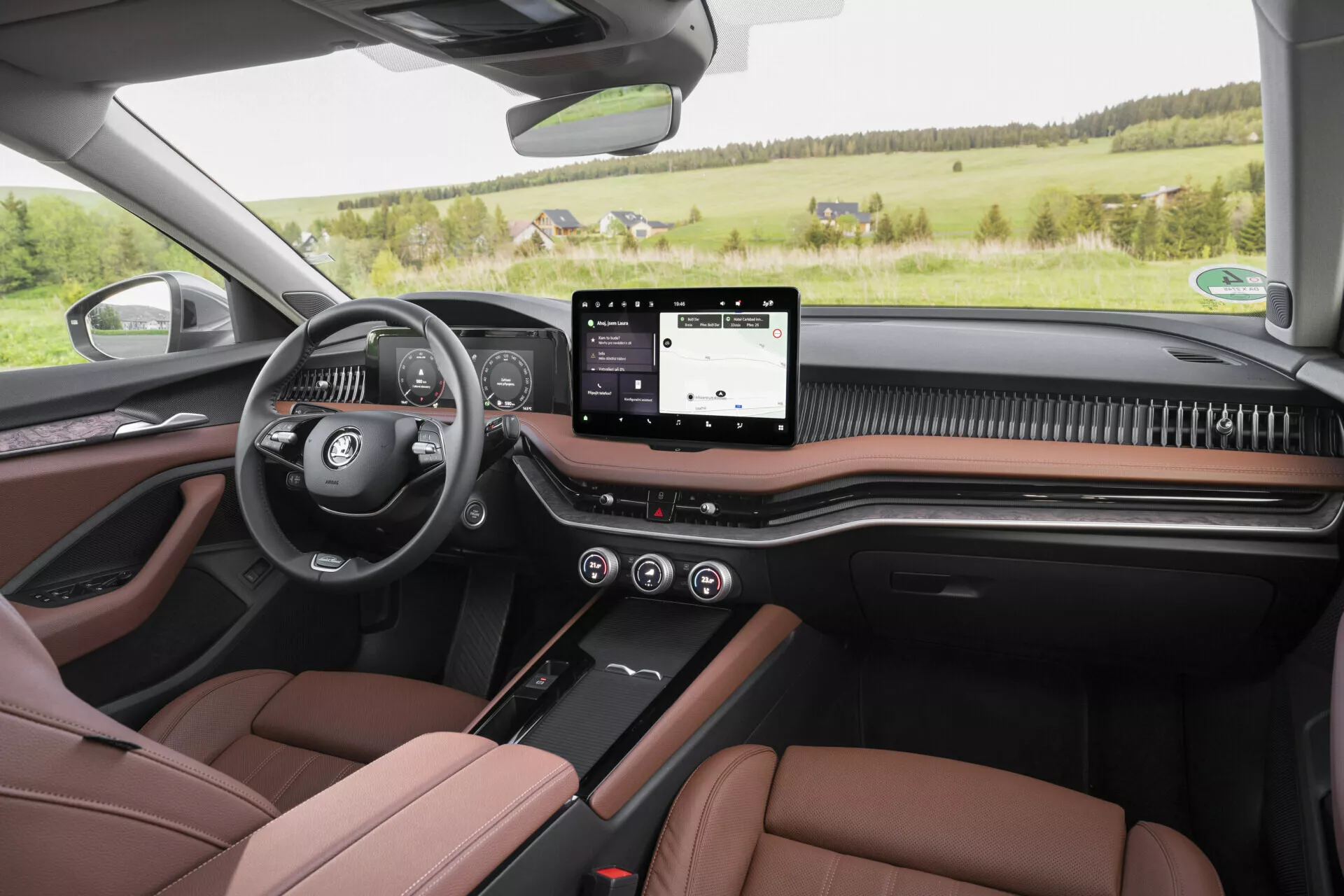 A first-person view from the rear seat of the 2024 Skoda Superb Limousine, showcasing a modern and luxurious interior with a left-side multi-function steering wheel, digital instrument panel, a wide touchscreen infotainment system, minimalistic climate and seat control buttons below, and a carbon fiber-like trim. The image also highlights two-tone brown and black leather front seats with contrast stitching, matching door panels with power controls, visible high-end audio system speakers, and a panoramic sunroof, all set against a rural landscape seen through the front windshield.