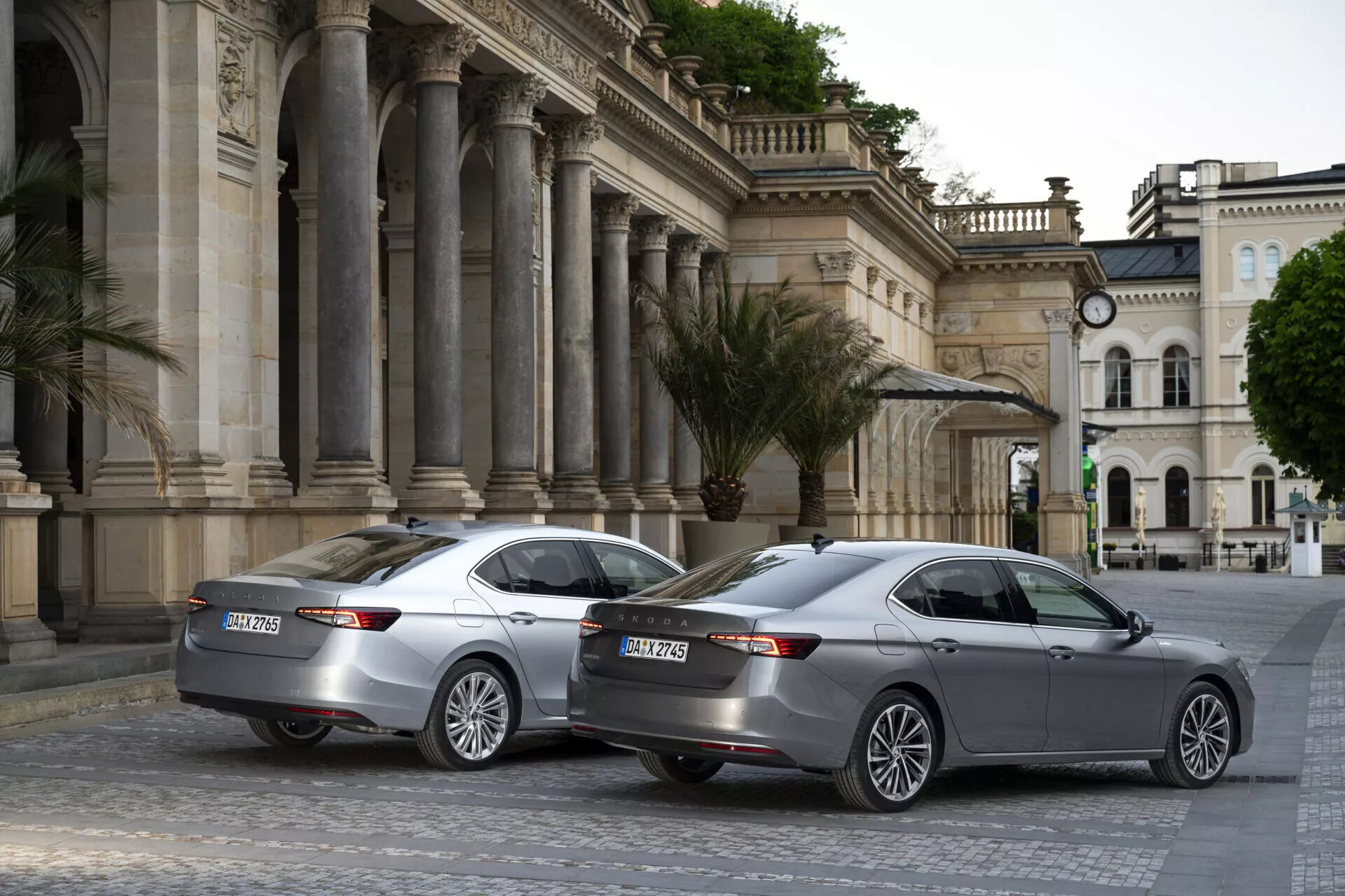 The image showcases two Skoda Superb 2024 sedans with identical models parked in an urban cultural center featuring classic architecture. Captured from a three-quarter rear-angle, the image highlights their sleek modern design with LED horizontal tail lights, dual exhaust outlets, parking sensors, and multi-spoke alloy wheels. Both cars have shark-fin antennas for advanced connectivity and a third brake light on the lower rear windshields. The scene is set under overcast or early morning/evening lighting conditions, adding a consistent subdued tone to the photograph.