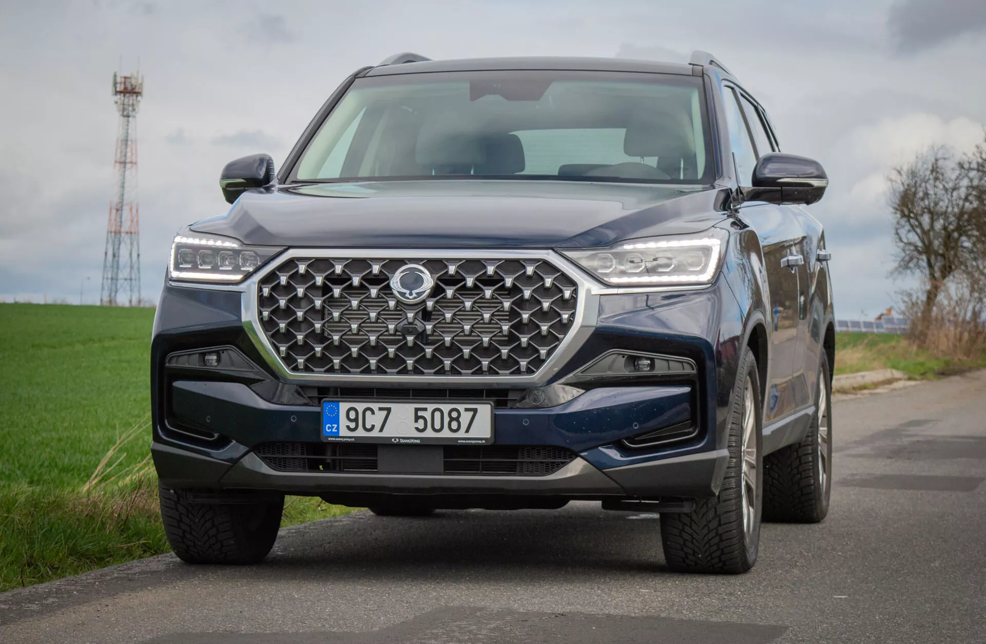 2021 SsangYong Rexton SUV on country road, showcasing a detailed view of its front-end design with a geometric grille and LED headlights, reflecting advanced technology and off-road capabilities