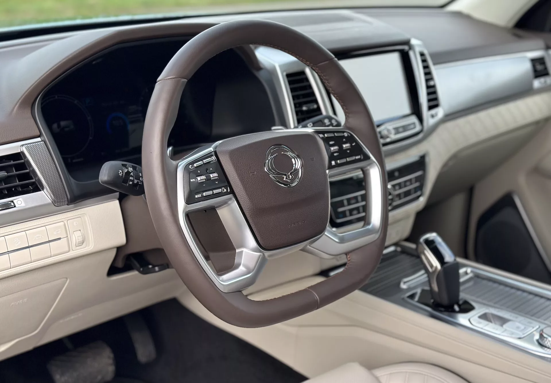 2021 SsangYong Rexton's luxurious interior view from the front passenger side, featuring a multifunctional leather-wrapped steering wheel with metallic accents, digital instrument cluster, advanced infotainment system, and a two-tone beige and dark color scheme.