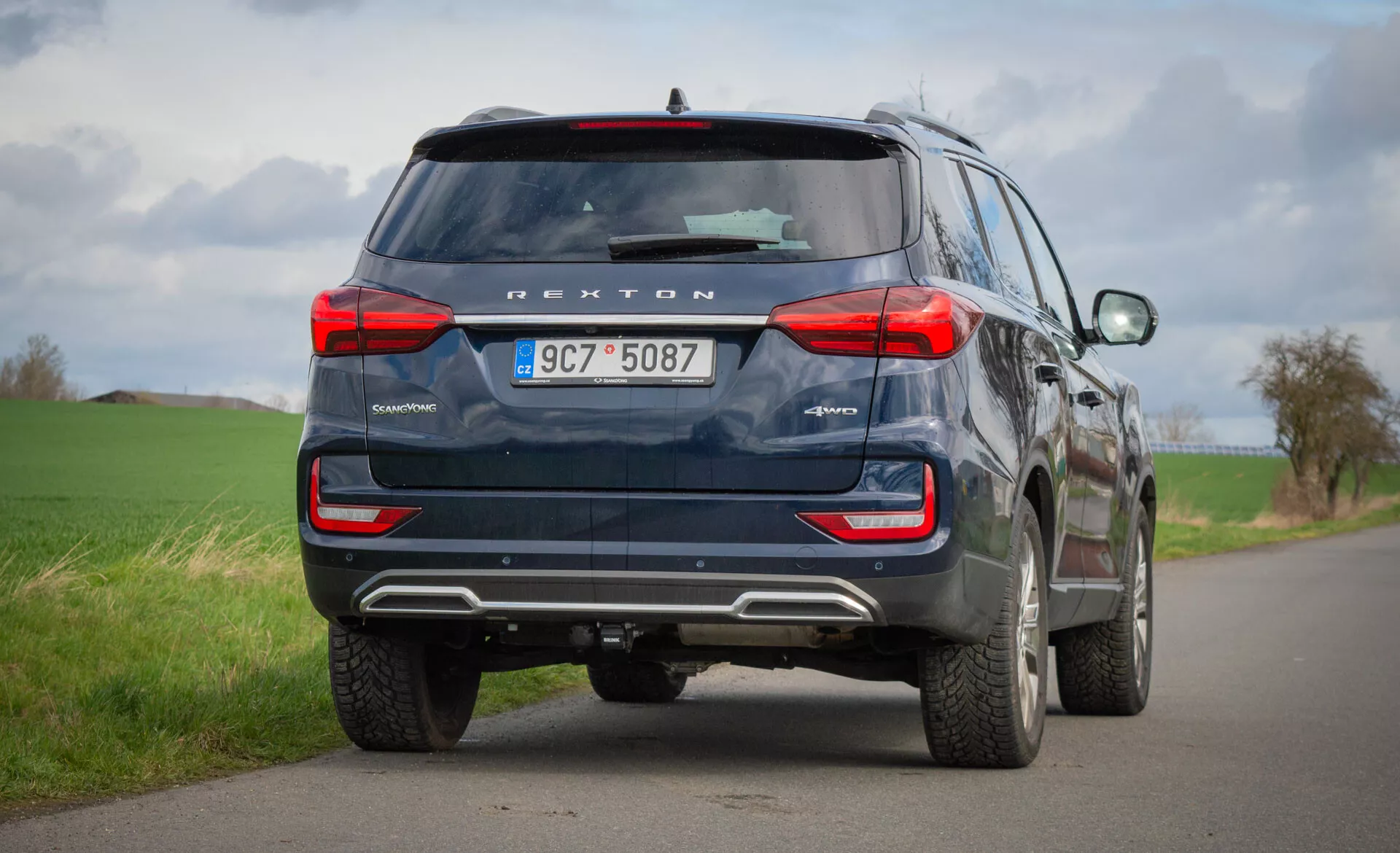 Rear view of a 2021 SsangYong Rexton blue SUV parked on a countryside two-lane road, highlighting its design features like taillights, rear window, exhaust pipes, and safety technology including rearview camera and parking sensors, under a cloudy sky.