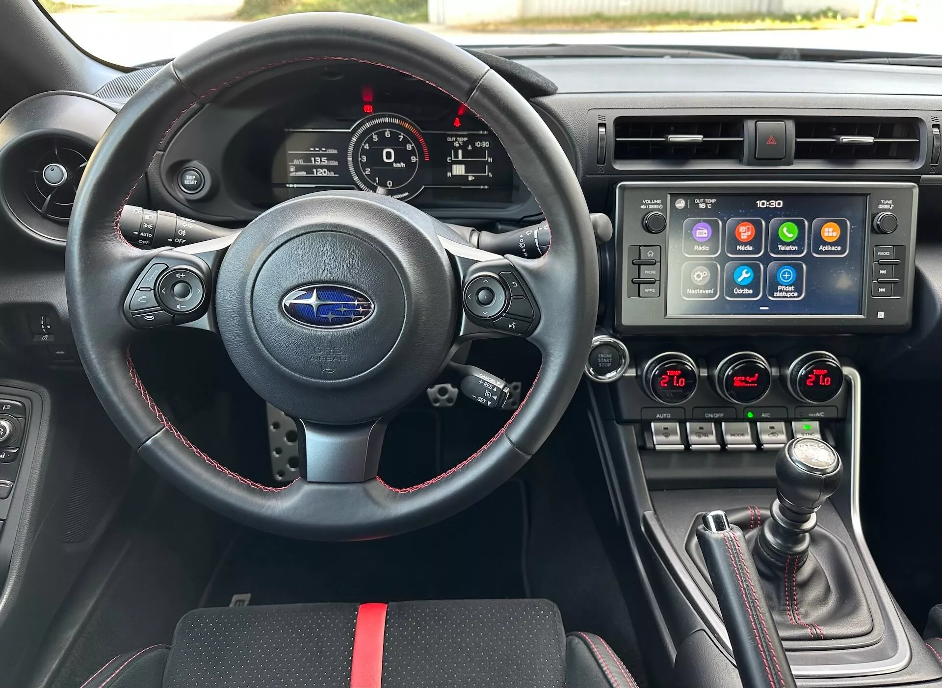 2023 Subaru BRZ showing interior with driver's perspective including a sporty leather steering wheel with multifunction controls and red stitching, an instrument cluster with analog dials and a digital display, a center console with a large touchscreen infotainment system and dual-zone climate control, and a manual gearshift with leather boot and red accents, against a black interior with sporty red accents, highlighting advanced vehicle features and technology.