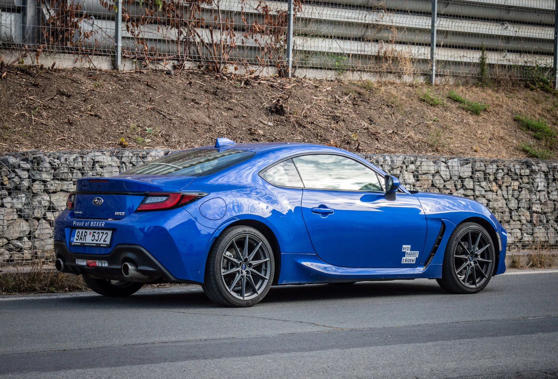 2023 Subaru BRZ bright blue sports coupe with aerodynamic design parked on a street, featuring a rear wheel arch, tail lights, rear window curvature to trunk lid, sculpted lines, sloping roofline, multi-spoke alloy wheels, performance brakes, low-profile rear spoiler, dual exhaust pipes in rear diffuser, body-colored side mirrors, and sporty long hood.