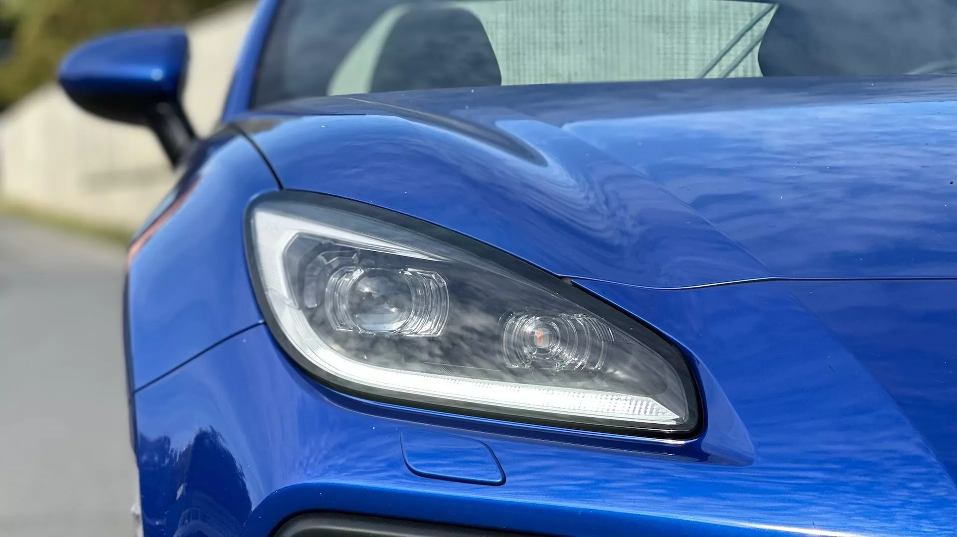 2023 Subaru BRZ close-up showcasing the driver's side headlight design, featuring LED or HID lighting, projector lenses, and a sporty air intake against a metallic blue bodywork
