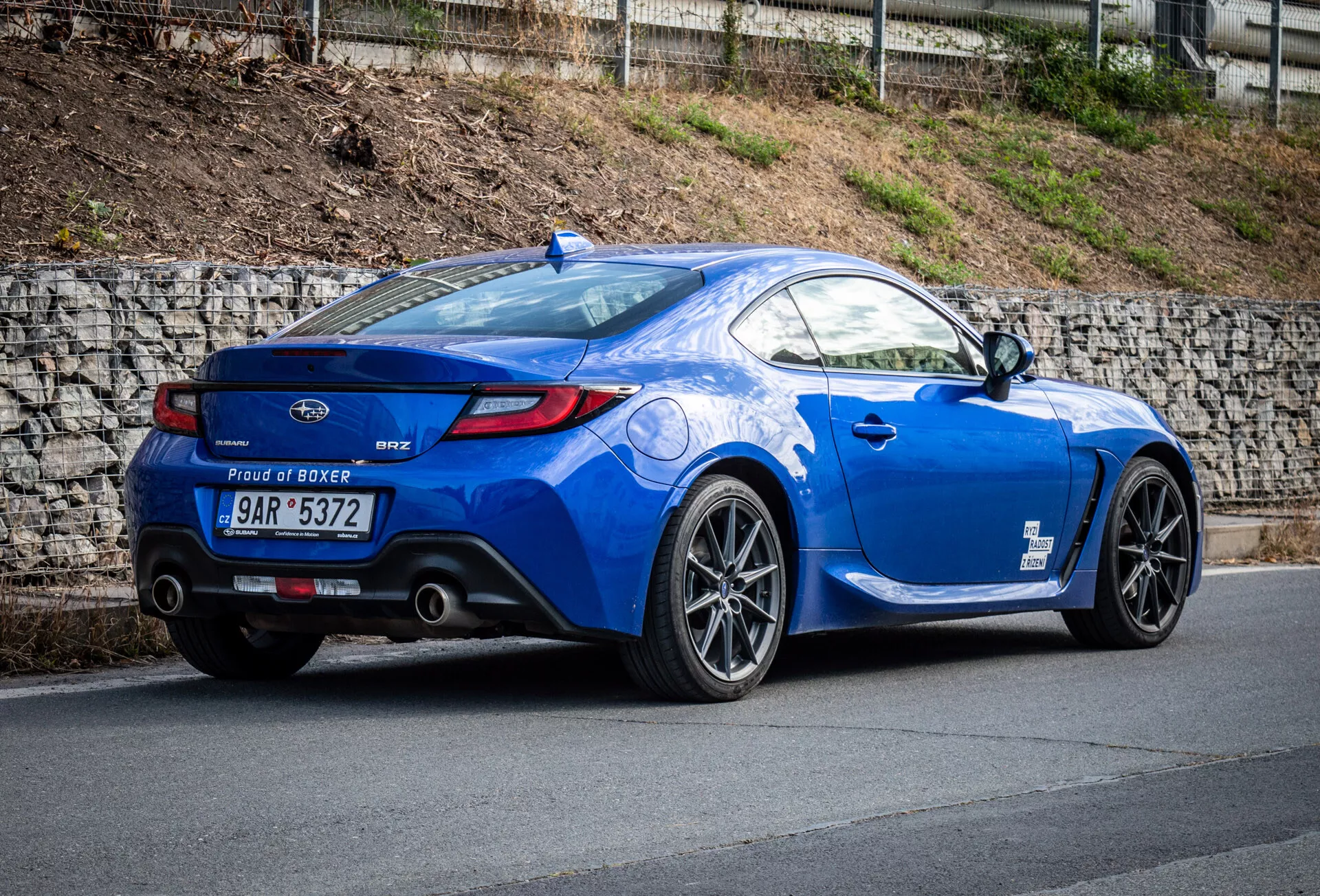 2023 Subaru BRZ sports coupe in metallic blue parked on an incline, showcasing its rear three-quarter view emphasizing the vehicle's side profile, curved rear, sporty alloy wheels, dual exhaust tips, lip spoiler, and 'Proud of Boxer' engine badge.