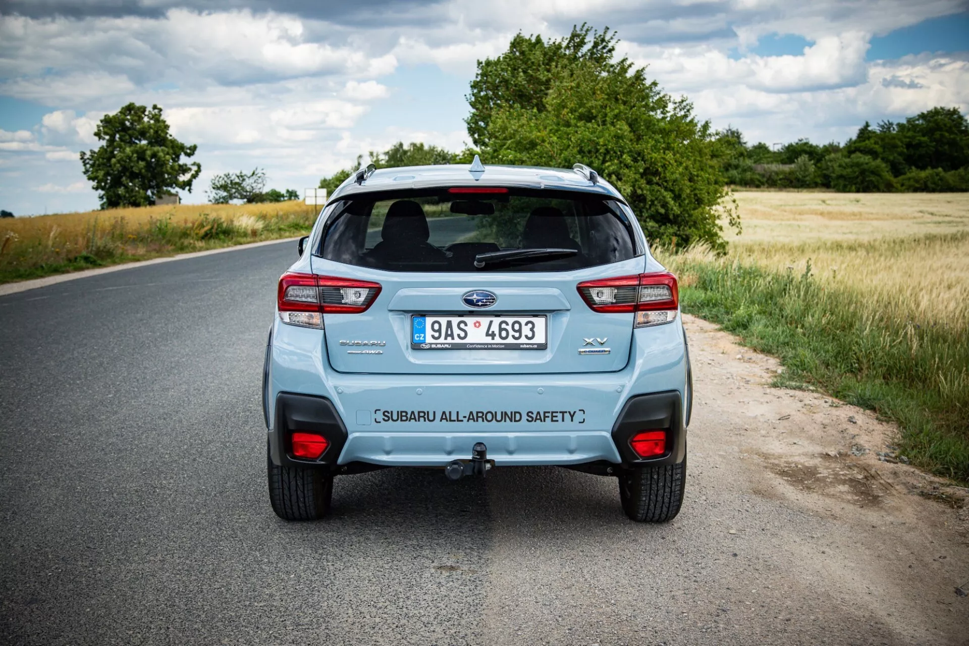 Rear view of a 2021 Subaru XV on a rural road, showing the car's distinctive taillights, dual exhaust system, and safety-tagged liftgate under partly cloudy skies