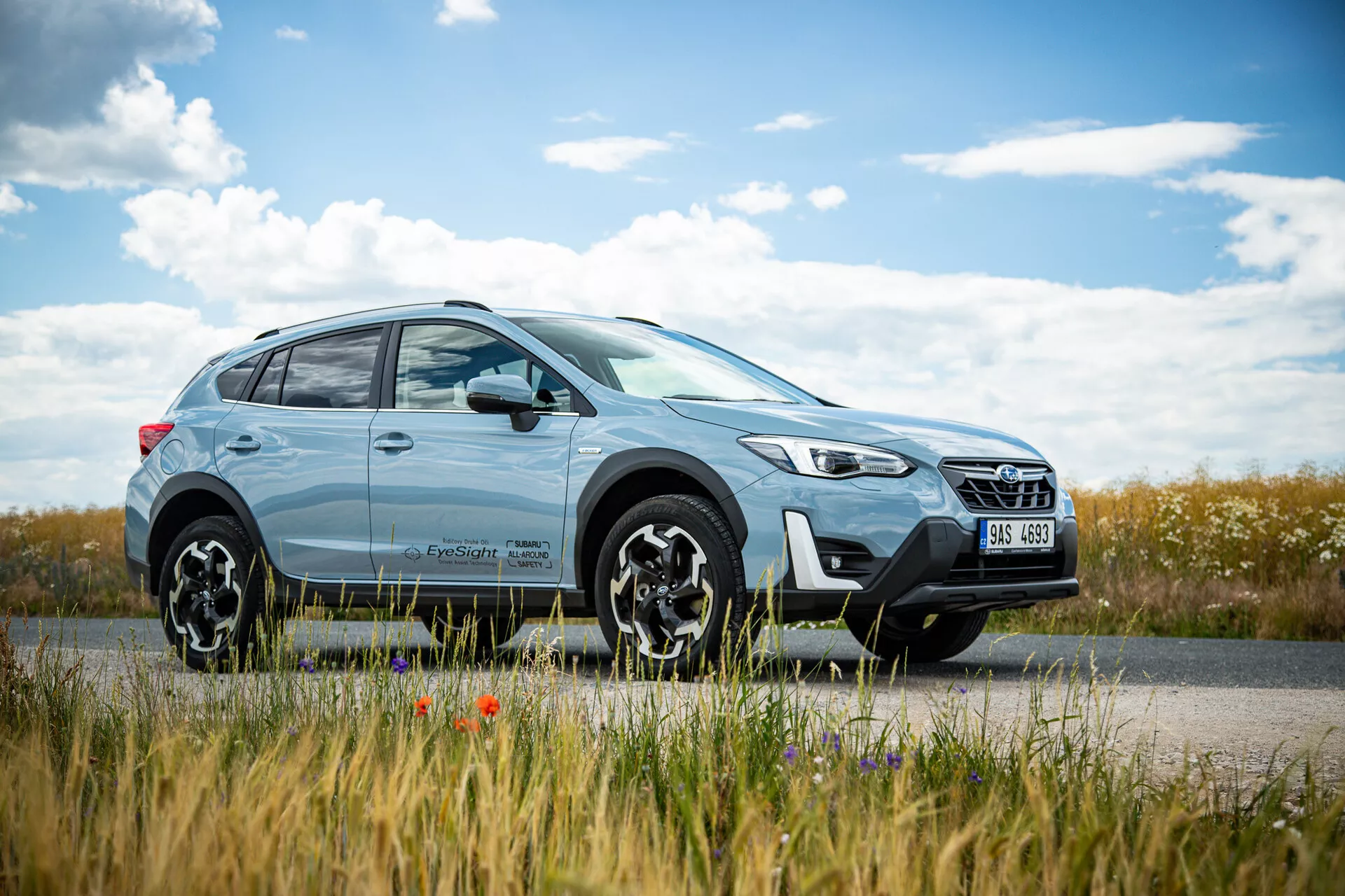 Light blue Subaru XV 2021 crossover SUV parked roadside with countryside landscape in the background, featuring modern design with LED headlights and multi-spoke alloy wheels, emphasizing both city and off-road capabilities.