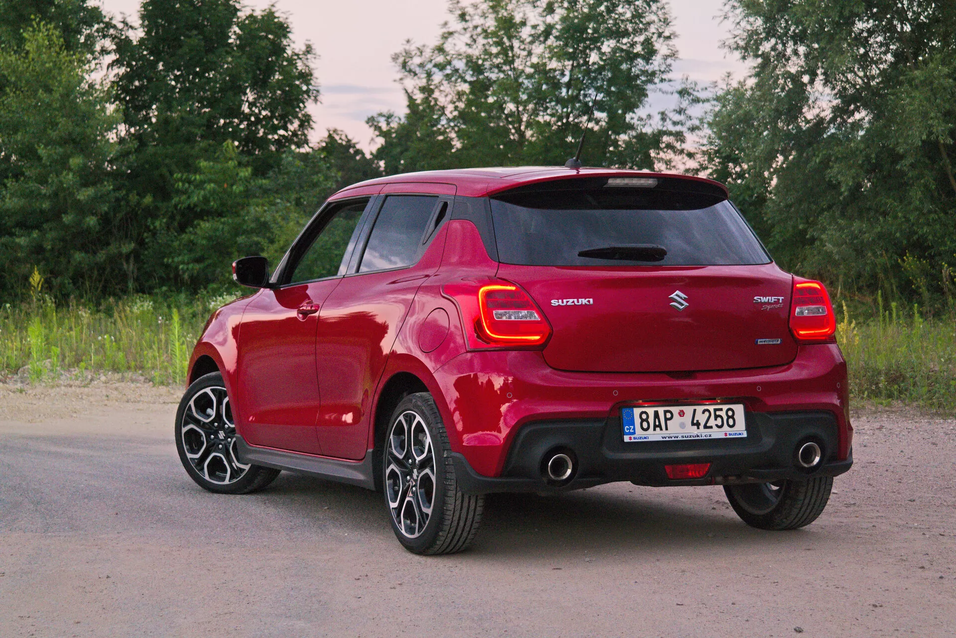 Image of a red Suzuki Swift 2017 Swift-Sport in a rear three-quarter view, showcasing the modern design and styling elements like LED taillights, integrated spoiler, shark-fin antenna, multi-spoke alloy wheels, body-colored door handles and side mirrors, and hybrid technology badging. The natural backdrop adds a serene and dynamic feel.