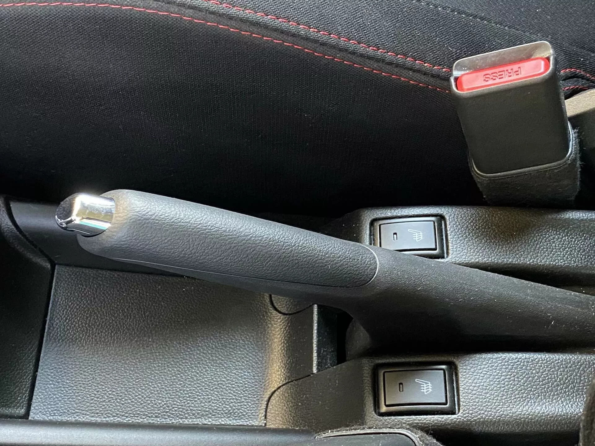 Close-up view of Suzuki Swift 2017 (Swift Sport) interior focusing on a leather-stitched handbrake lever with a metallic button and seat heater control buttons. The handbrake lever is centrally positioned and slightly angled left, indicating it is engaged. Two push-button switches with LED indicators and a seat icon, located next to the handbrake, suggest dual-zone heating for front seats. A seatbelt buckle with a red release button is partially visible behind the controls. The image is taken from an elevated angle with natural daylight lighting, highlighting the technology and comfort-focused features.