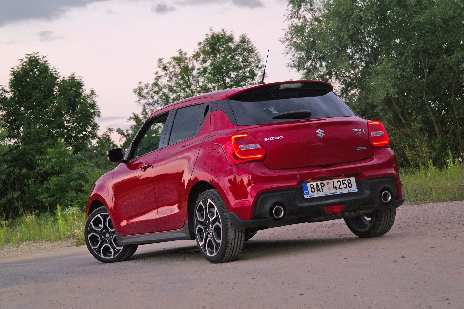 Red Suzuki Swift Sport 2017 hatchback captured from rear three-quarter angle, highlighting its back and driver side profile. The photo, taken from a slightly lowered perspective, emphasizes the modern multi-spoke alloy wheels and low-profile tires. Dawn or dusk lighting with partly cloudy conditions provides soft natural light. Visible features include contemporary taillight design with LED elements, manufacturer's badge, model name, body-colored side mirrors possibly with turn signal indicators, rear bumper with integrated parking sensors, and a roofline hinting at a spoiler for aerodynamic efficiency.