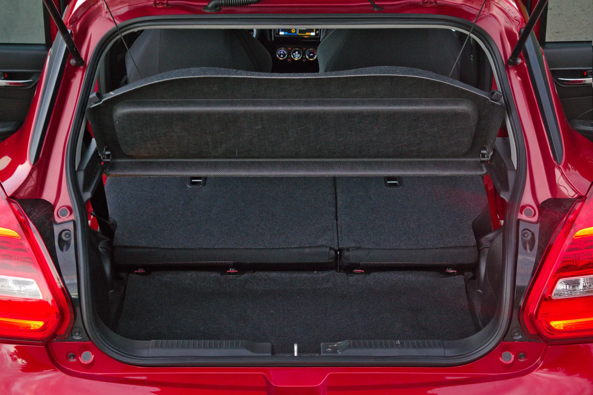 Rear trunk space of a 2017 Suzuki Swift Sport hatchback with open hatchback door, showcasing empty cargo area with dark automotive carpet. View from direct rear perspective, revealing removable and foldable parcel shelf, and 60/40 split rear seats. Partial view of rear windshield wiper and interior components including rear speaker and lighting system.