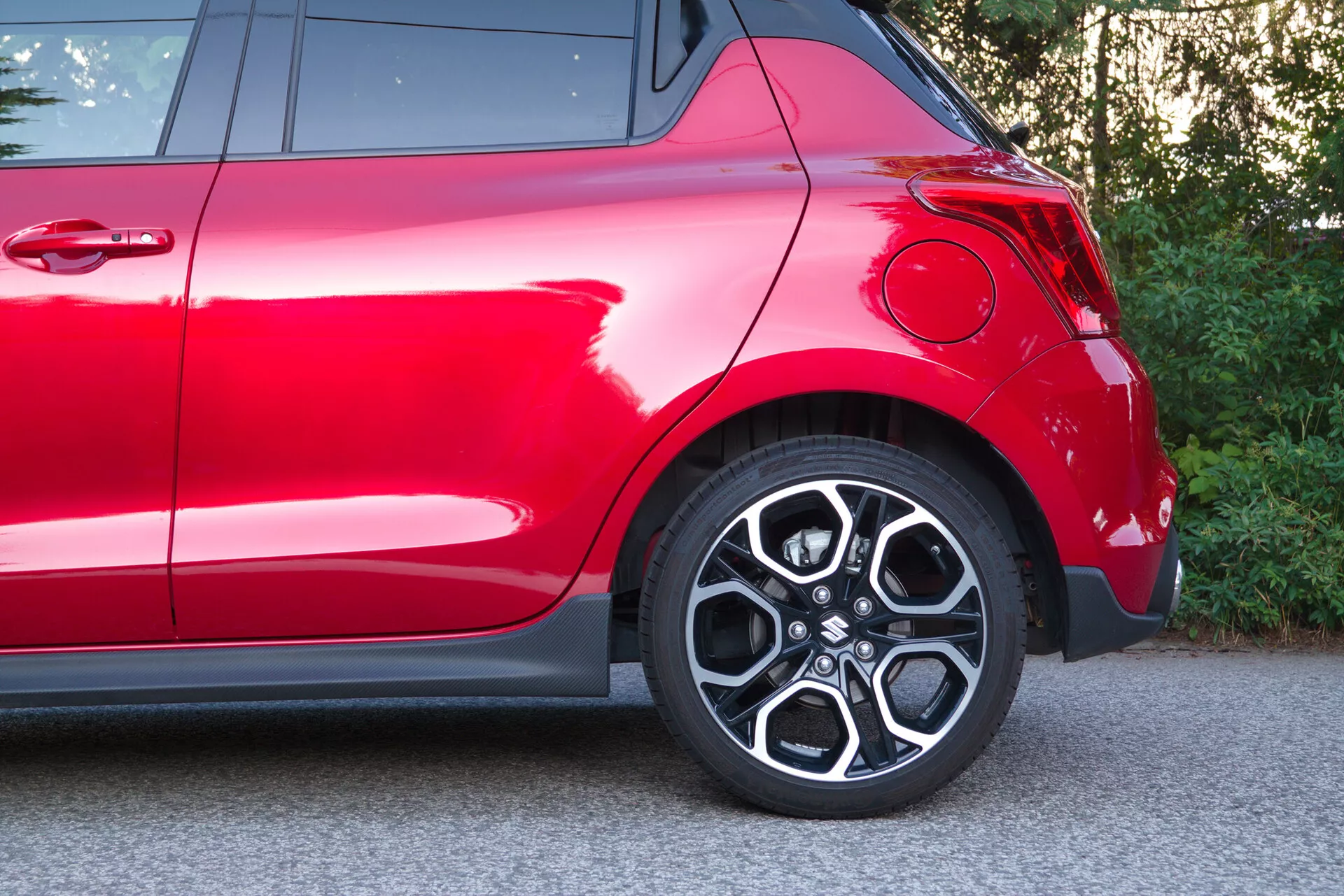 Partial side view of a red Suzuki Swift 2017 (Swift Sport), focusing on the rear quarter including the rear wheel and fender area. The close-up captures the stylish alloy wheel with a multi-spoke design, low-profile tire, modern curved rear fender, deep glossy red paint, part of the rear taillight, and chrome trim elements, highlighting its sporty design and premium exterior finish.