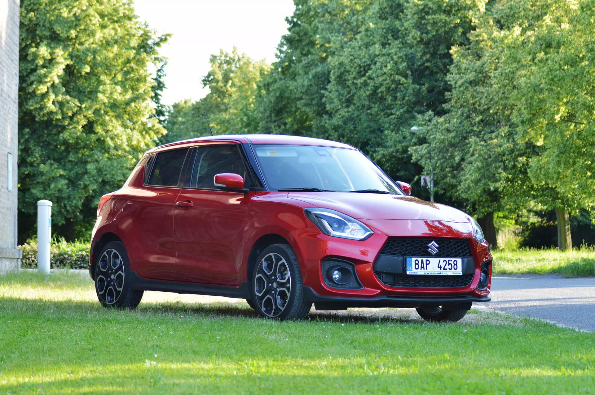 The image shows a red 2017 Suzuki Swift Sport hatchback positioned on a grassy area, likely in a park or roadside turf, captured from a front three-quarter angle. The prominent front grille, sleek and angular headlights, fog light assembly, and sloping hood into an aerodynamic windshield are highlighted. The alloy wheels, body-colored side mirrors, gently sloping roofline to the rear hatch window, and tinted windows contribute to the vehicle's design. The natural lighting and unobstructed view emphasize the car's overall stance and design.