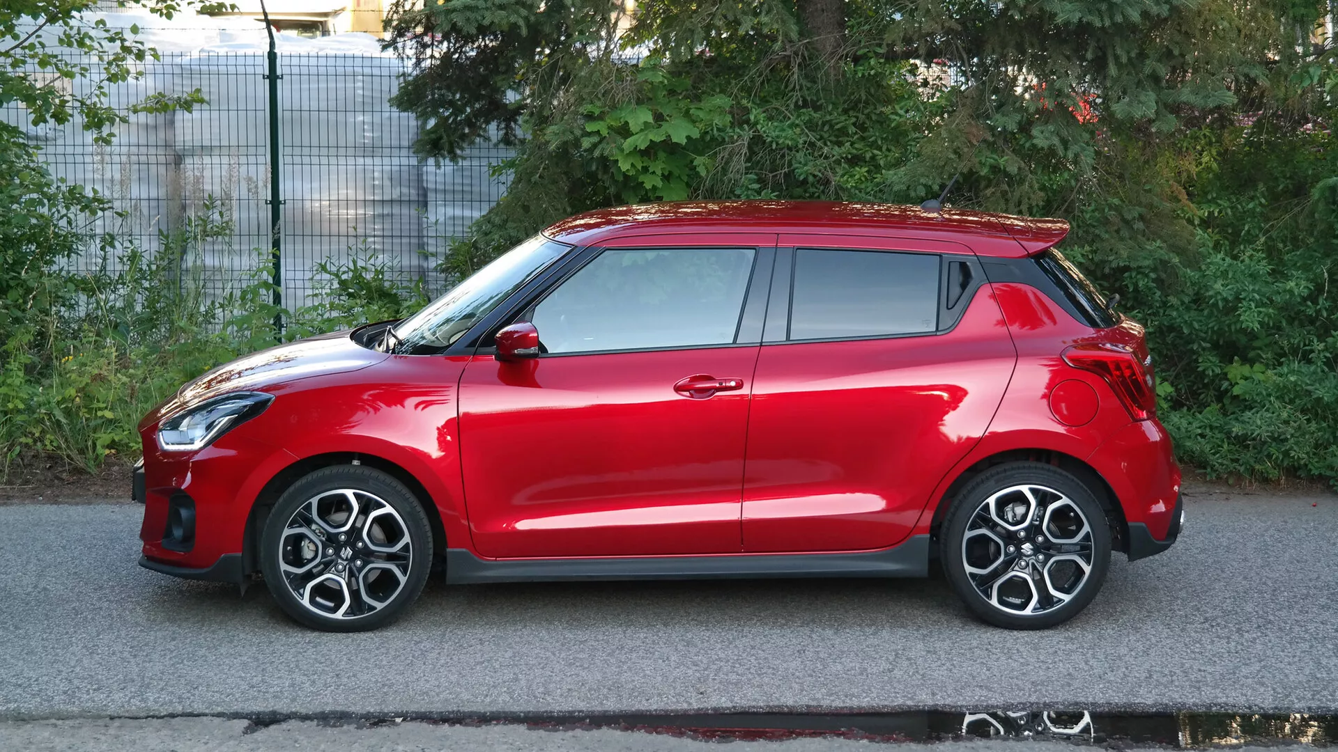 Side profile view of a red 2017 Suzuki Swift Sport hatchback car. The vehicle is captured in natural daytime lighting, showcasing its sleek design features, including headlight integration into the front wheel arch, multi-spoke alloy wheels, body-colored side mirrors and door handles, a remote keyless entry system, and a visible sunroof. The car also features a roof spoiler and high-mounted taillights. The image, set on a suburban street, emphasizes the car's aerodynamics, sporty characteristics, and likely advanced safety technologies.