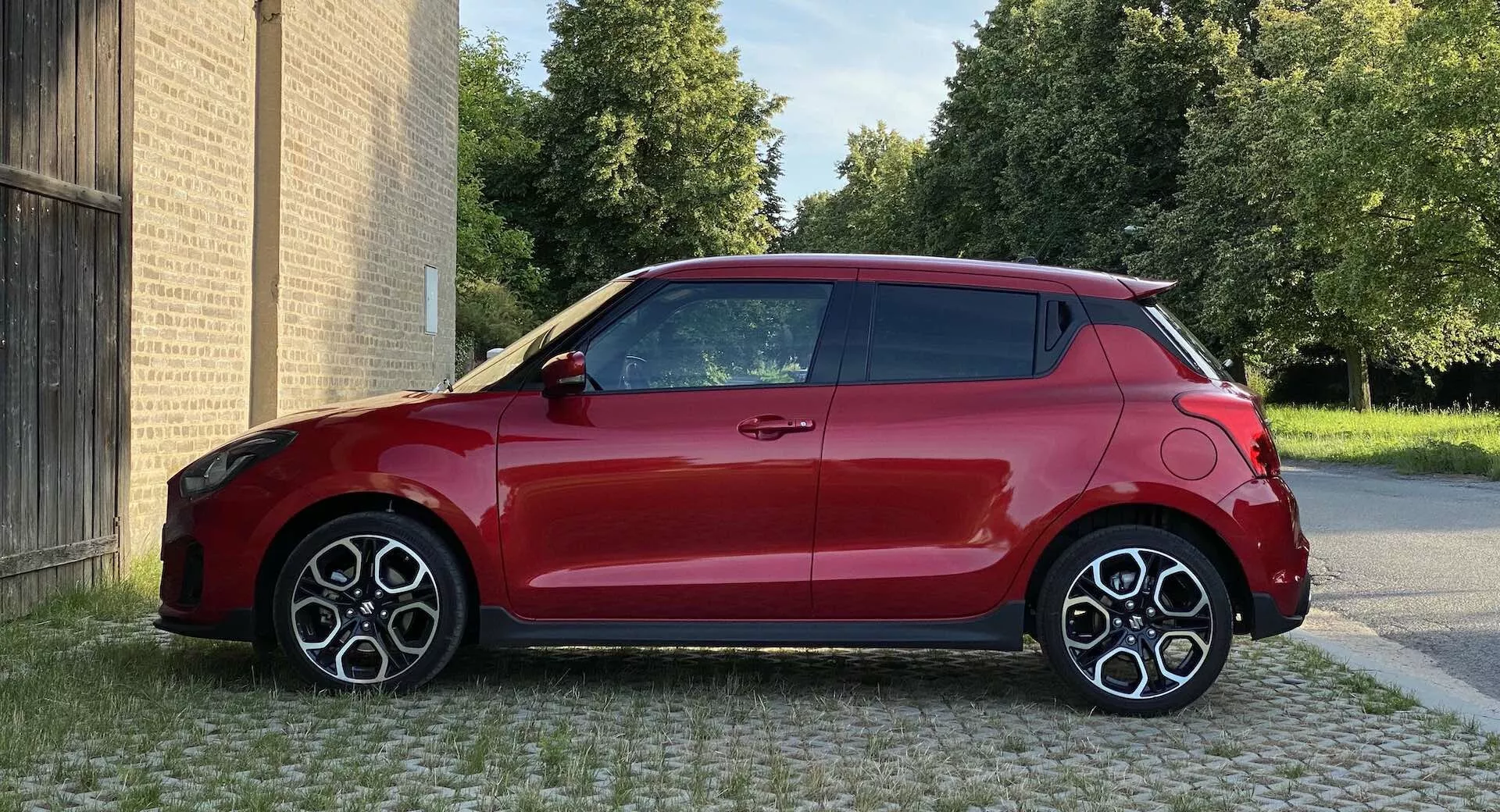 Image of a red Suzuki Swift Sport 2017 hatchback, taken from a side profile angle at eye level, showcasing the right side with a two-tone design of red body and black roof. The car is parked on a paved area with buildings in the background and trees visible behind the buildings. The compact, modern design features large sporty multi-spoke alloy wheels, visible aerodynamic lines, and a shark-fin antenna. The lighting is daylight, with sharp image quality and surface reflections discernible.