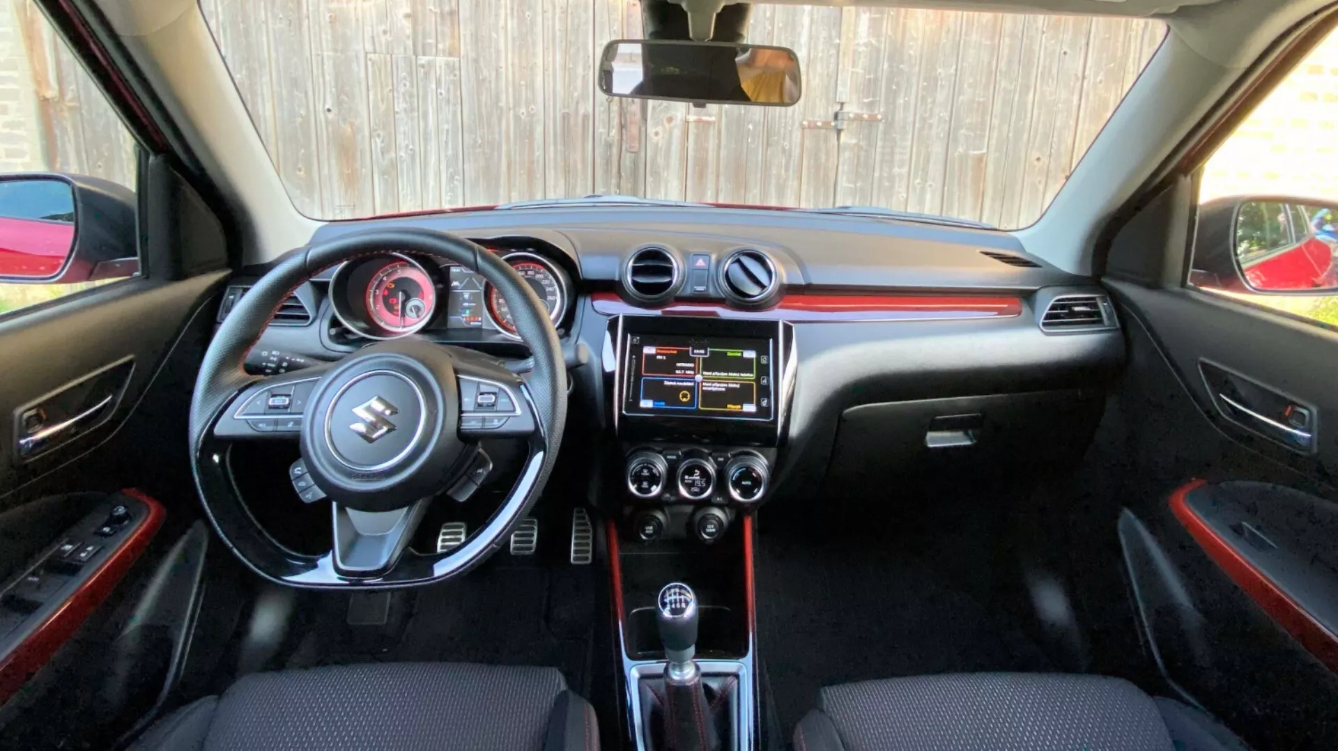 Interior shot of a Suzuki Swift 2017 (Swift Sport) from rear seat perspective looking towards front cabin, featuring a detailed view of the dashboard, steering wheel with mounted controls, central console with infotainment system and climate controls, automatic transmission gearshift, and black interior materials with red accents.