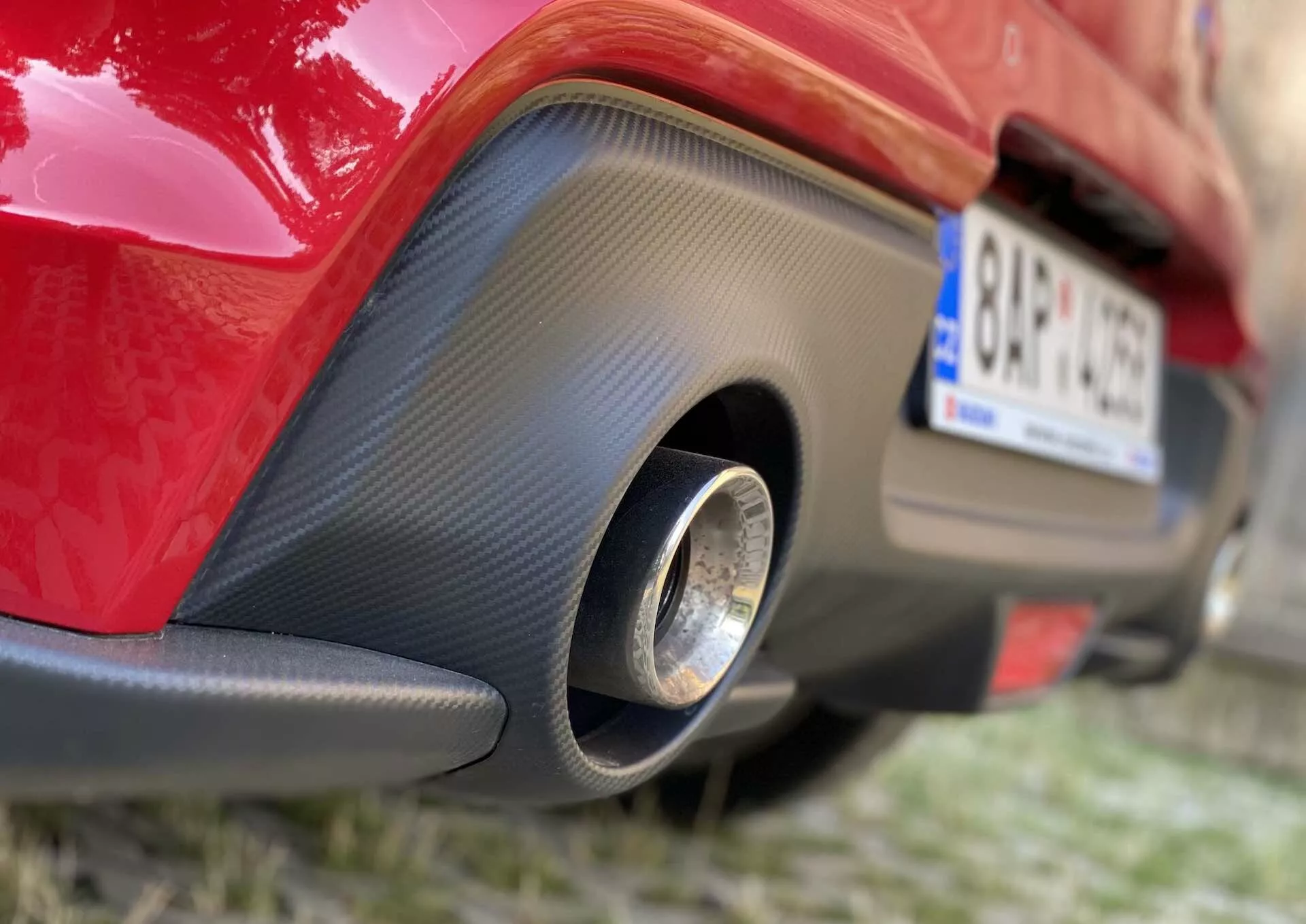 Close-up shot of the rear section of a red 2017 Suzuki Swift Sport, focusing on the lower part of the rear bumper. The photo captures a low-angle view from the right, highlighting a black carbon fiber-like insert with a single round metallic exhaust pipe at the center.