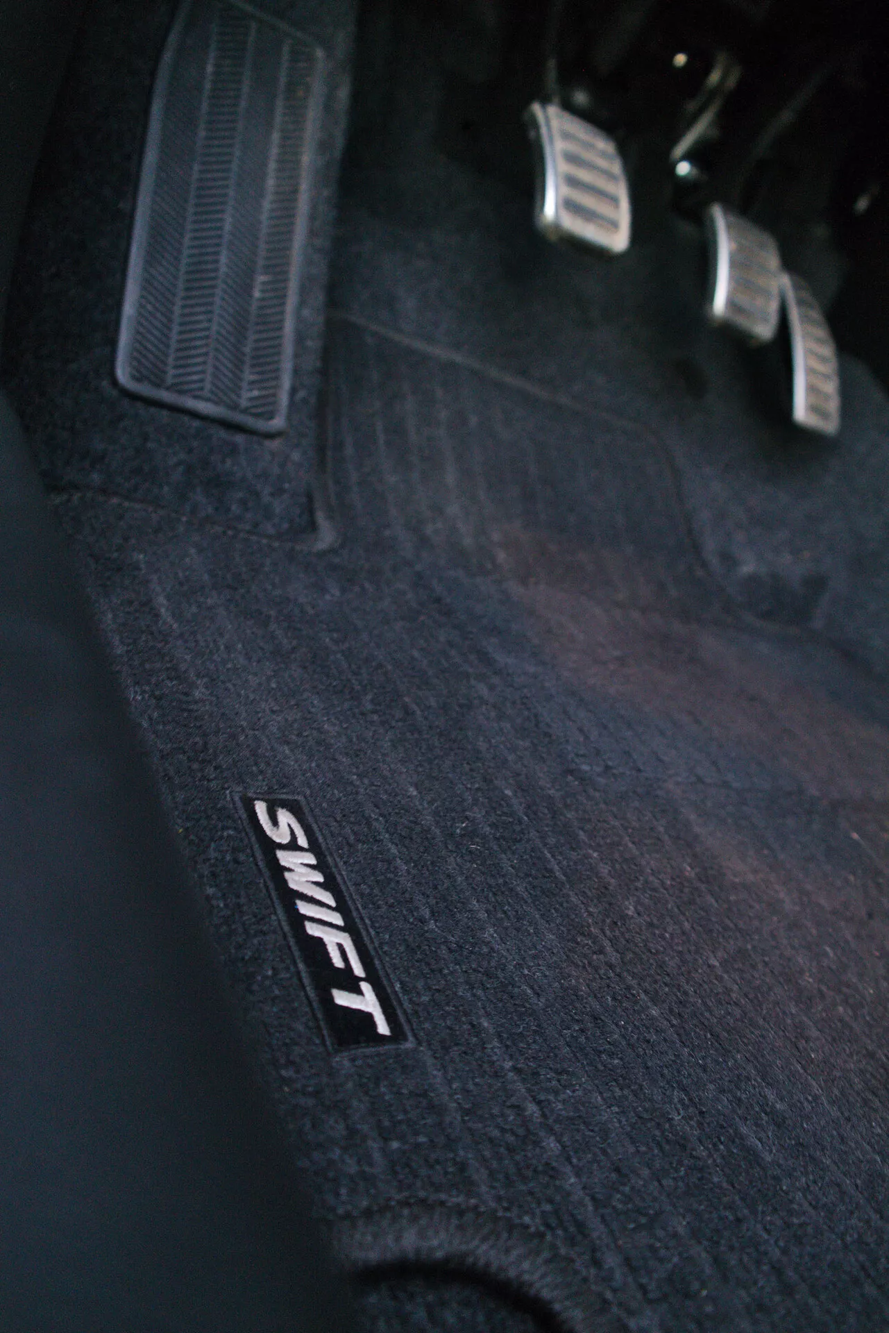 Close-up view of the driver's footwell area in a 2017 Suzuki Swift Sport, showcasing the interior details. The image prominently features a carpeted floor mat with the stylized branding 'SWIFT' visible. It highlights the vehicle's arrangement of three pedals designed for a manual transmission, with metal surfaces and black rubber anti-slip inserts: clutch on the far left, brake in the middle, and accelerator on the right. The angled perspective captures the textured design and ergonomic features, including a corner of the driver's footrest.