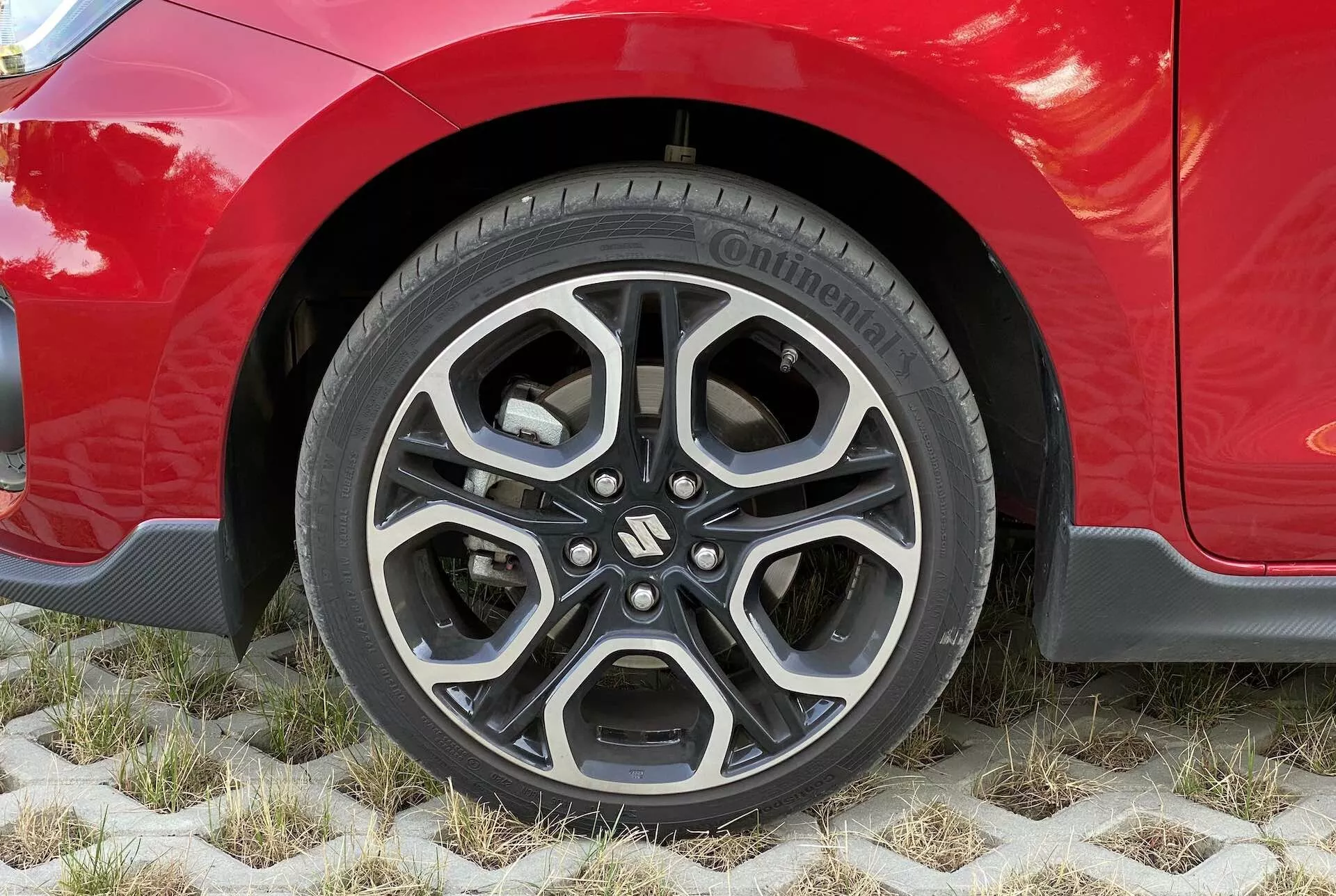 Close-up of a red Suzuki Swift 2017 Swift Sport's wheel, showcasing a modern alloy rim with a complex, multi-spoke design in black and machined silver finishes. Visible tire is a Continental PremiumContact, indicating high-quality and good tire condition. The car's shiny, well-maintained bodywork extends above the wheel, highlighting the fender's aesthetic lines and possibly sporty trim. The vehicle is parked on eco-friendly hexagonal pavers allowing grass growth, emphasizing sustainable parking solutions.