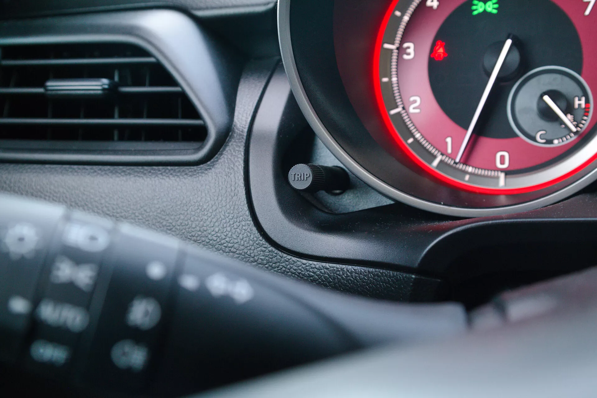 Interior view of a 2017 Suzuki Swift Sport focusing on the instrument cluster and part of the steering wheel from the driver's perspective. The image highlights the tachometer with red accents, a 'TRIP' button, and a glimpse of another gauge, possibly the temperature gauge. Multi-function controls on the steering wheel suggest integrated audio and cruise control settings.