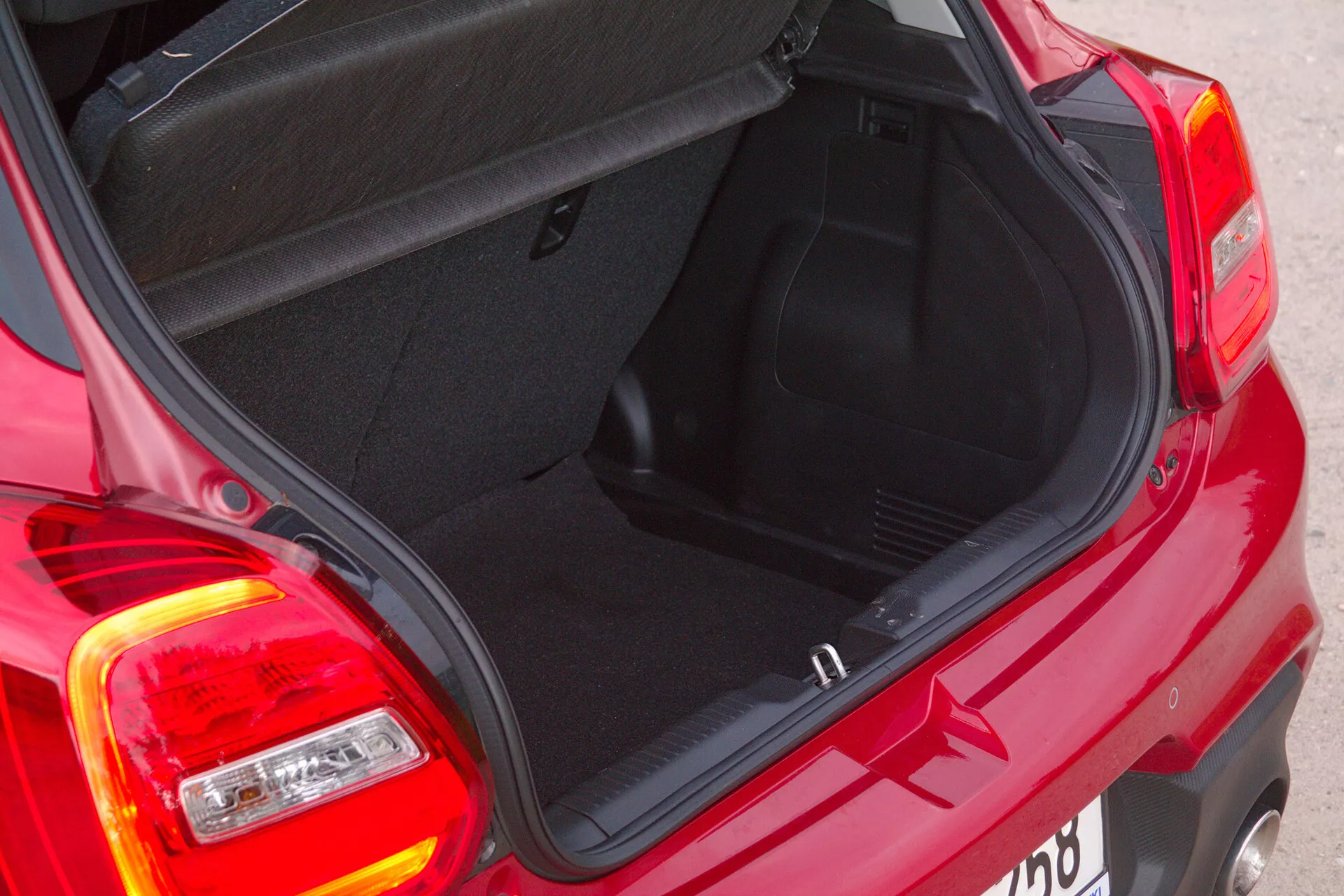 Close-up, high-angle view of the rear of a red 2017 Suzuki Swift Sport, focusing on the open trunk area. The shot is taken from the right-rear quarter, showcasing the trunk interior, trunk lid, and design of the compact car's rear lights. The brake lights are under a red cover, while reversing lights and turn indicators are under a clear cover. The trunk reveals standard features like visible latches and hinges, and the carpeting on the bottom and sides. The low load lip suggests easy cargo handling.