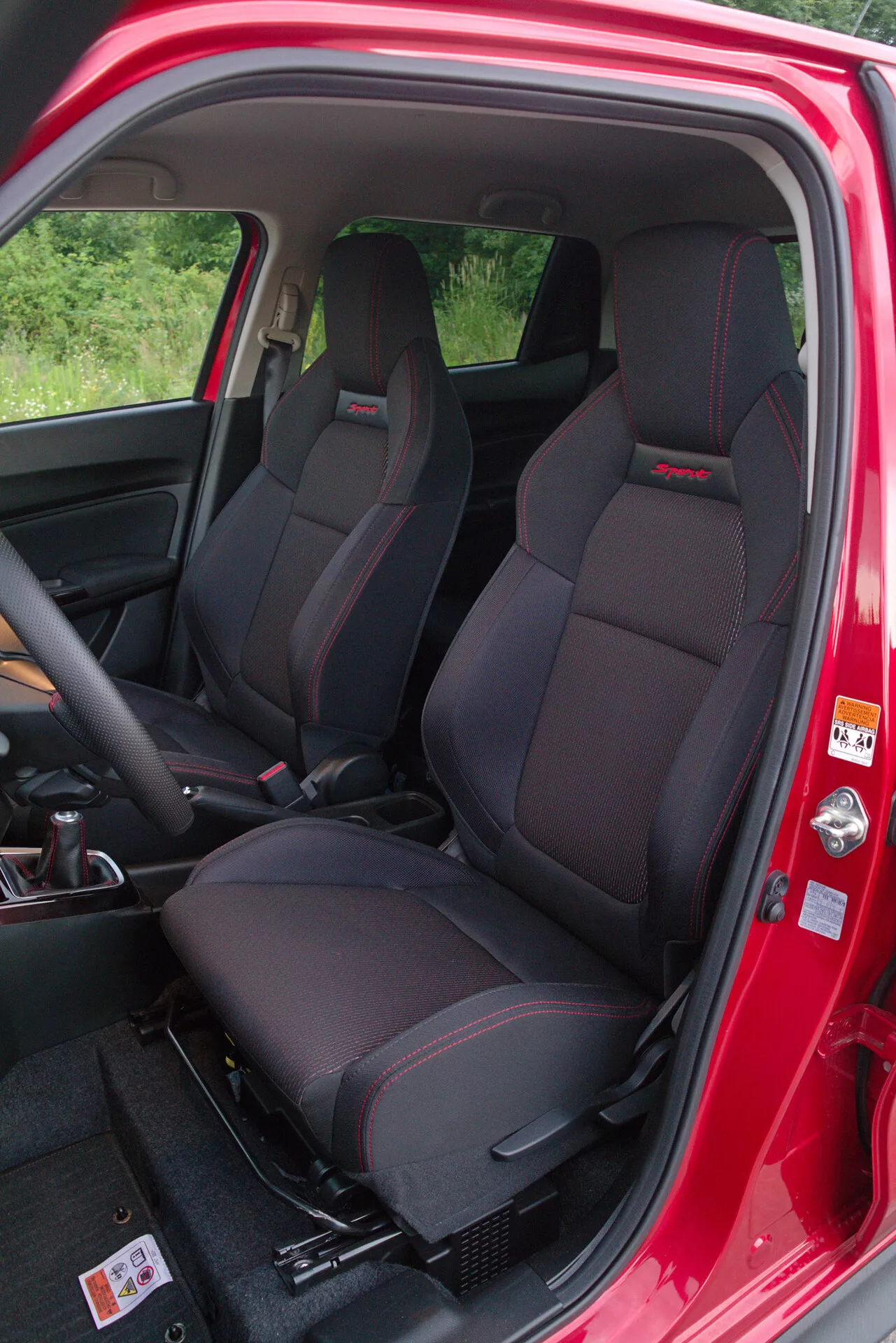 Interior view of the 2017 Suzuki Swift Sport from the passenger side, highlighting front bucket seats with integrated headrests and red stitching, open passenger door with red exterior paint and warning label, visible interior door panel with controls and door pocket, partial view of dashboard and steering wheel, and dark carpeted flooring with durable plastic sill.