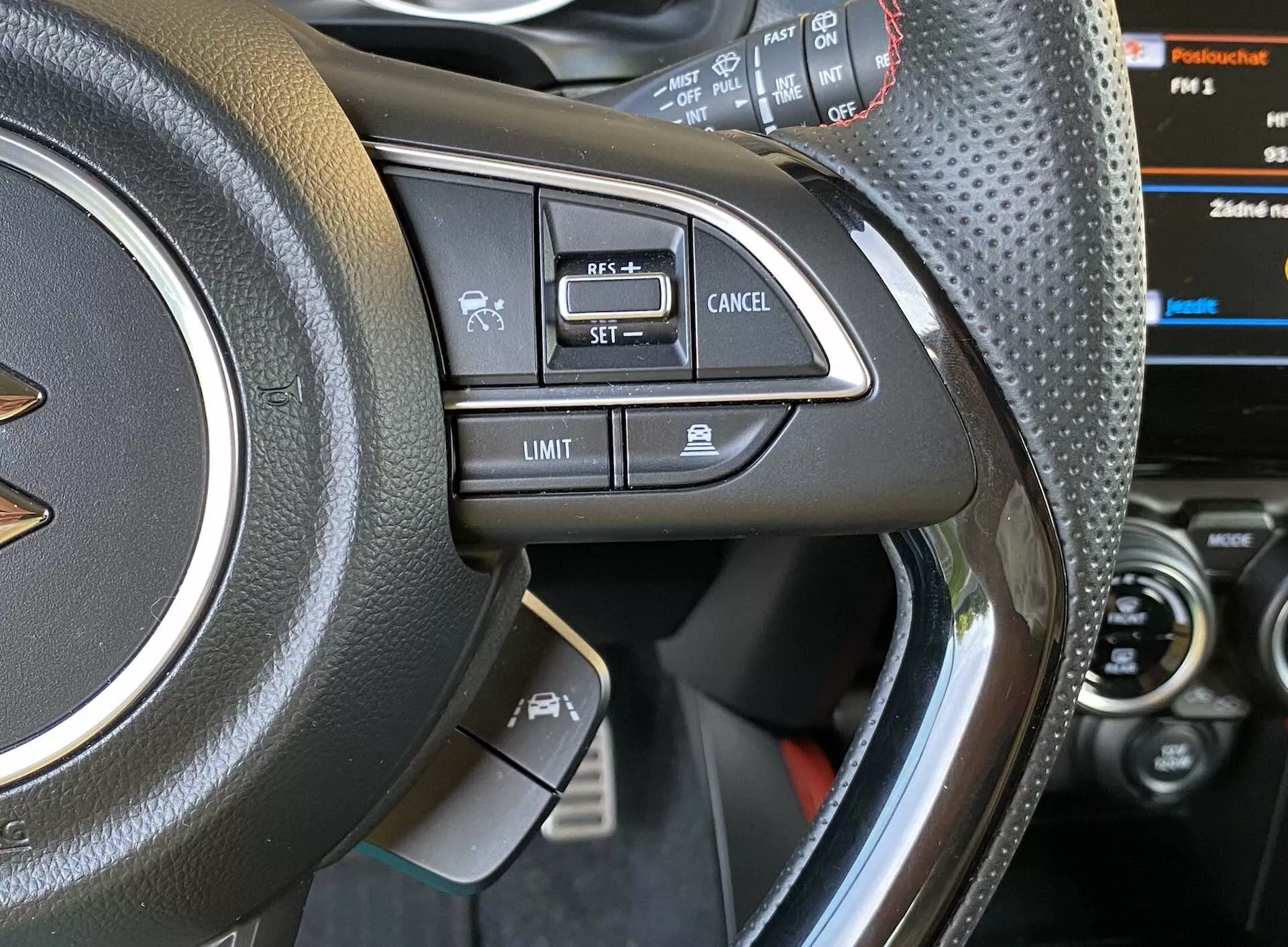 Close-up view of a Suzuki Swift 2017 Swift-Sport's left side of the steering wheel, showcasing mounted controls. Photo taken from the driver's seat or slightly outside the driver-side door, focusing on leather or leather-like material with stitching, metallic accent, and various buttons like voice command, Bluetooth hands-free calling, cruise control 'SET,' 'Cancel,' and speed limiter 'Limit.' Partially visible instrument cluster with digital display and car's center console.