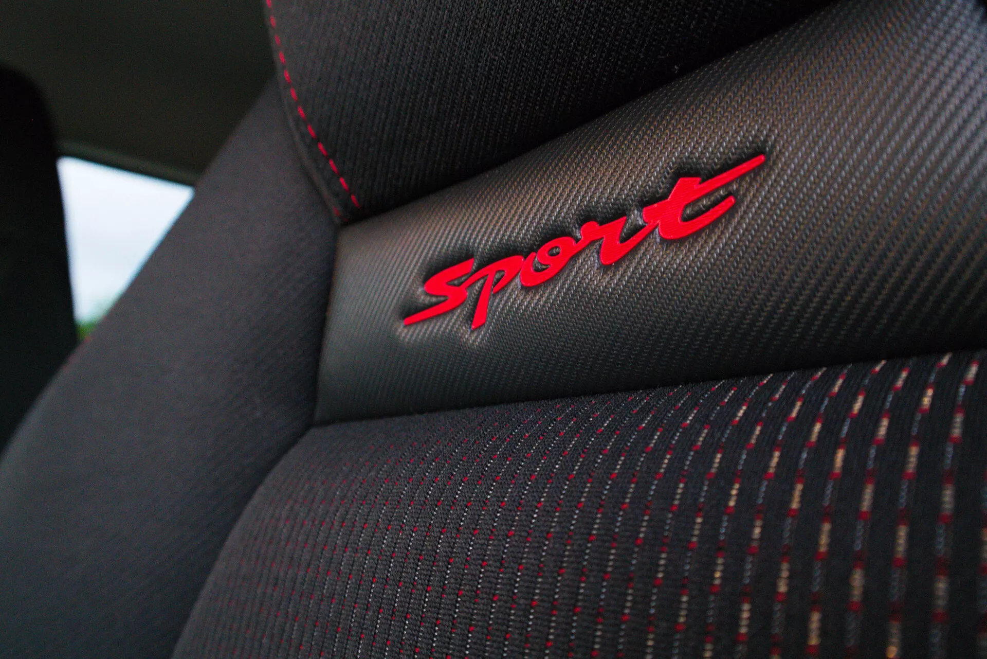 Close-up shot of a Suzuki Swift Sport 2017 car seat's backrest, showcasing detailed fabric texture, red stitches, and 'Sport' embroidery on a black fabric, emphasizing the vehicle's sporty trim level.