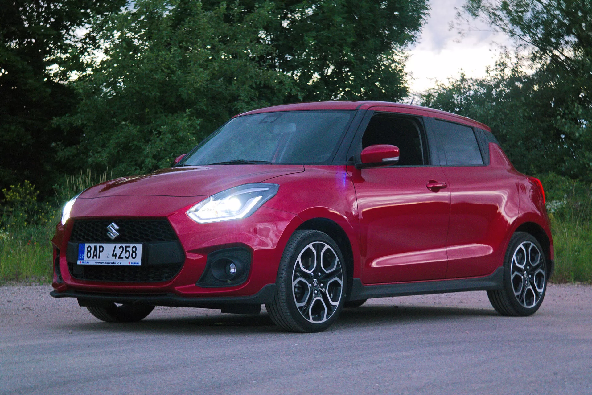Three-quarter front view of a red hatchback car, showcasing modern design elements such as a prominent front grille, sleek angular headlights likely featuring LED technology, multi-spoke alloy wheels, and a dynamic contour line along the side profile. The vehicle has body-colored door handles and integrated turn signals on the side mirrors. The glossy red paint finish is even and free of damage, parked on an asphalt surface with a backdrop of greenery, indicating a rural or suburban setting.
