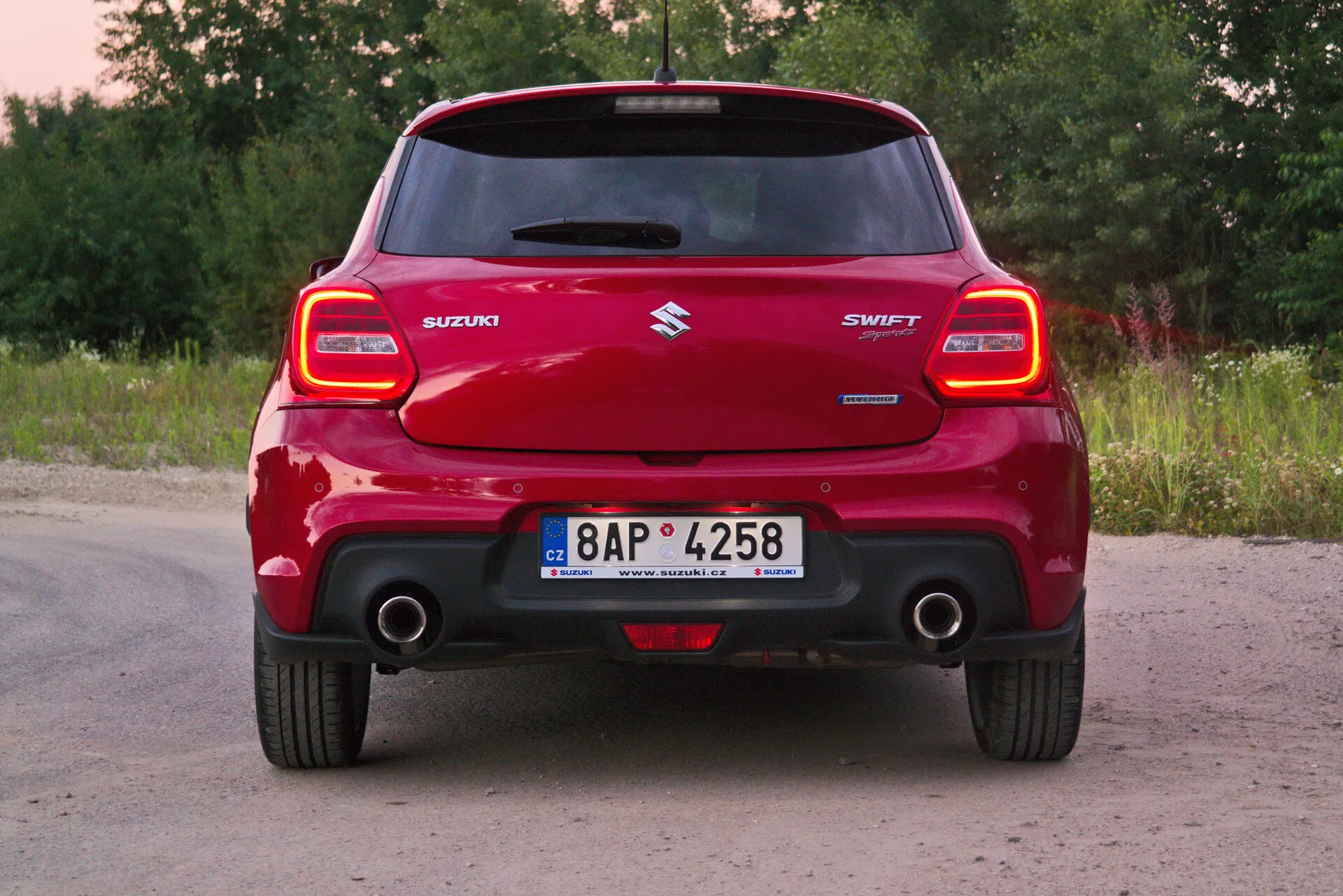 Rear view of a red 2017 Suzuki Swift Sport car, focusing on its full width and prominent rear bumper, distinctive rear light clusters, roof spoiler, central Suzuki logo, 'SWIFT' model name, visible exhaust pipe, and registration plate in a rural setting with greenery and trees.