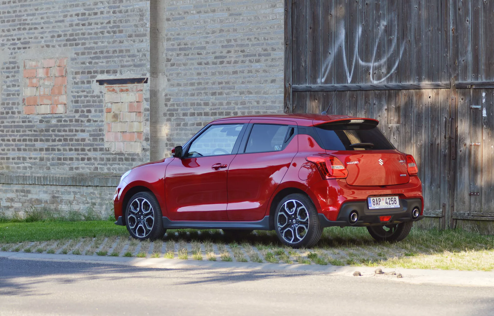 2017 Suzuki Swift Sport hatchback in bright red, captured from a three-quarter rear view, parked against an old building backdrop. The image highlights design features such as tail lights, rear hatch, prominent rear spoiler, dual-tone bumper with diffuser, single exhaust tip, multi-spoke alloy wheels, fully painted body including side mirrors and door handles, indicating a higher trim level or luxury options.