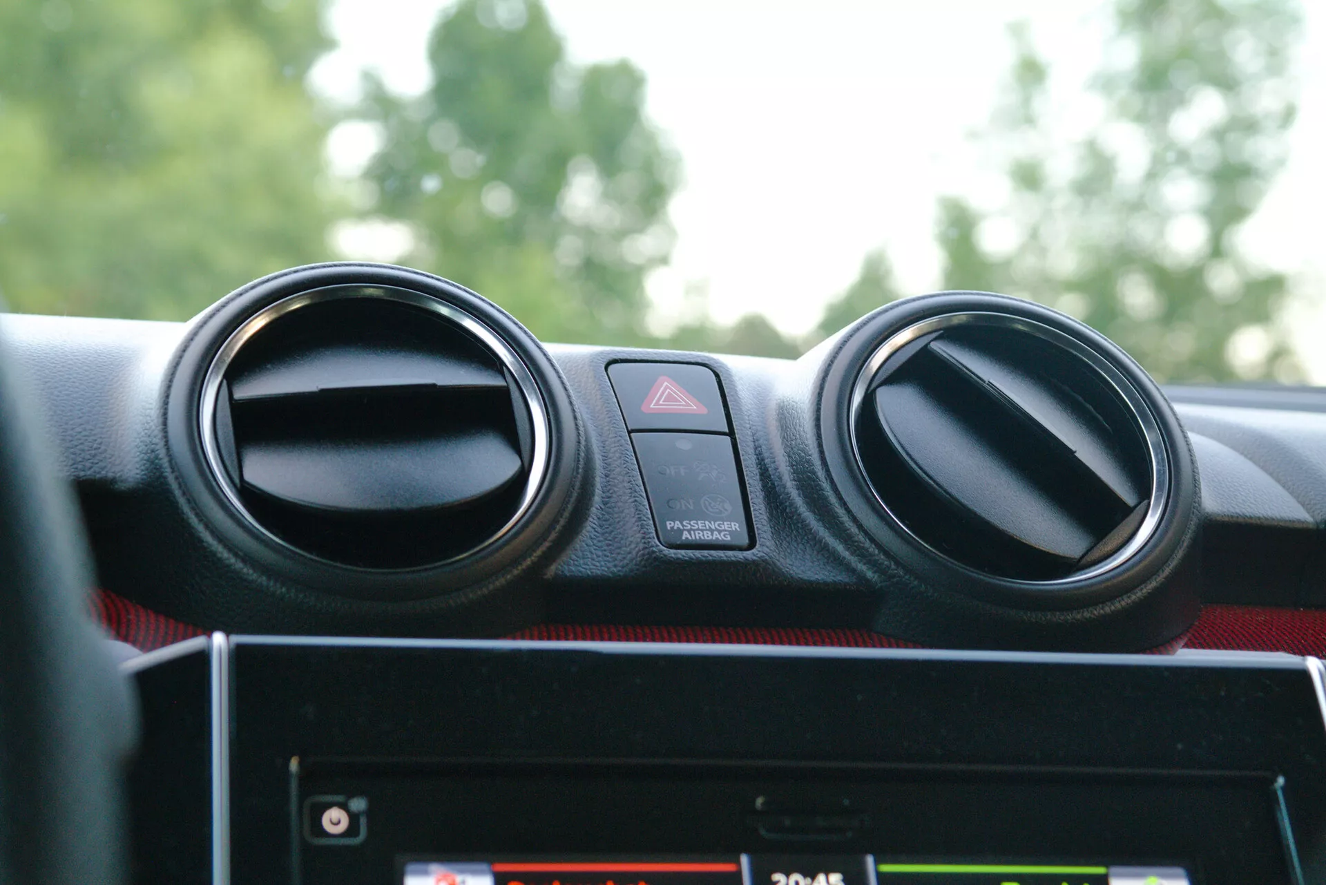 Close-up of the 2017 Suzuki Swift Sport's interior, focusing on the center console dashboard. The image reveals two circular metallic-finished air conditioning vents, a centrally-placed hazard warning button with a red triangle symbol, and a partially obscured infotainment system below a top-opening compartment. The dashboard has a soft texture with red accents, indicating a sporty or premium trim. Lighting suggests a daytime setting with natural light reflecting off the right air vent, emphasizing the modern, ergonomic design and user-friendly interface.