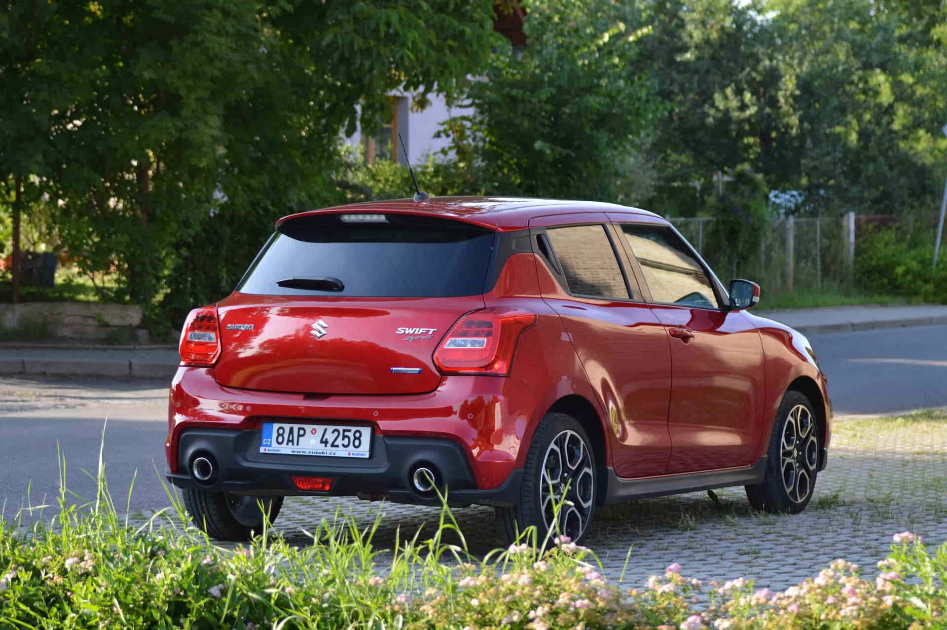 The image features a vibrant red Suzuki Swift Sport 2017 hatchback captured from a rear three-quarter angle, showcasing the side and back. Highlights include a contrasting black roof and pillars, a raised rear spoiler, distinct tail lights, and a model badge 'Swift'. The photograph is taken at a level height, displaying five-spoke alloy wheels in motion, suggesting the car is being driven. The lighting indicates it’s early morning or late afternoon, casting long shadows. The car’s technological features, albeit not entirely visible, likely include rear parking sensors and a rearview camera.