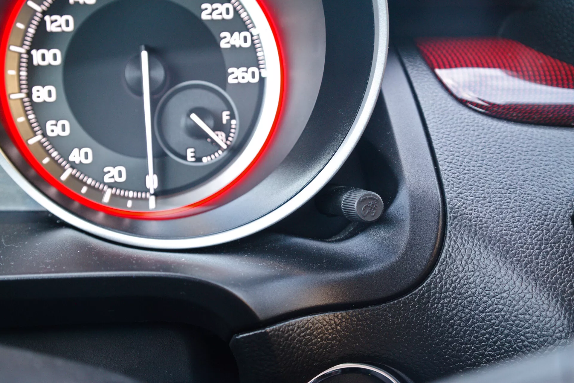 Close-up view of the instrument cluster of a 2017 Suzuki Swift Sport, highlighting the speedometer and fuel gauge. The speedometer has white numerals in increments of 20, reaching up to 260 km/h or mph, with the needle at zero. The design features a sporty red and white theme with a red circle on the inner dial circumference. The nearly full fuel gauge is to the right. The shot is angled from the driver's seat with a black granulated texture around the gauges and a color-matched red and black door interior, potentially leather and plastic. A small circular object is near the speedometer.
