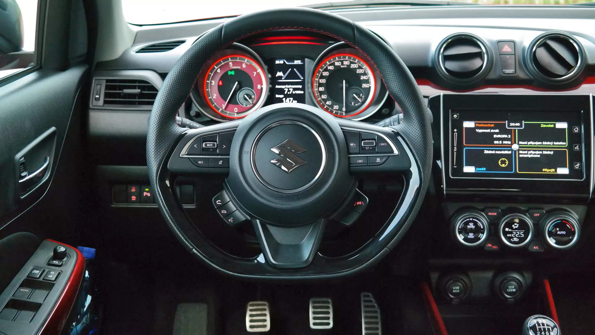 Interior shot of a Suzuki Swift Sport 2017 from the driver's perspective, featuring a three-spoke flat-bottom steering wheel with red stitching and control buttons, analog tachometer and speedometer with a central digital display, a touchscreen infotainment system, dual-zone climate control with red accent lighting, circular silver-accented air vents, and cup holders in the center console.