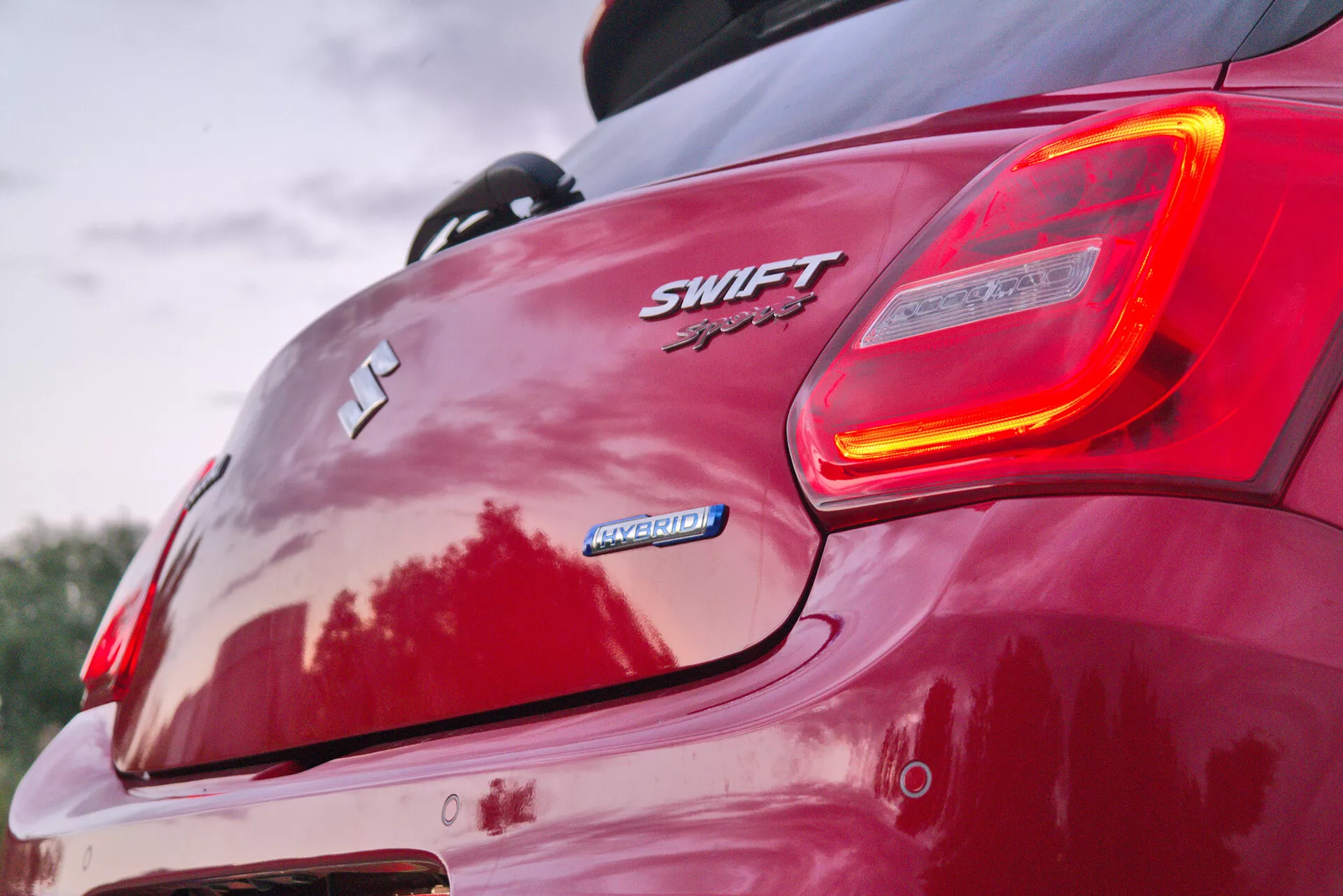 This close-up image captures the rear, right-hand corner of a red 2017 Suzuki Swift Sport, focusing closely on the car's badging and illuminated taillight. From a low rear angle, it accentuates the car's rear contours, spoiler, and sporty aesthetics. Visible features include the Suzuki 'S' emblem, 'SWIFT' and 'Sport' badges, 'HYBRID' badge, the design of the rear bumper, alloy wheels, and reflections that suggest a glossy paint finish.