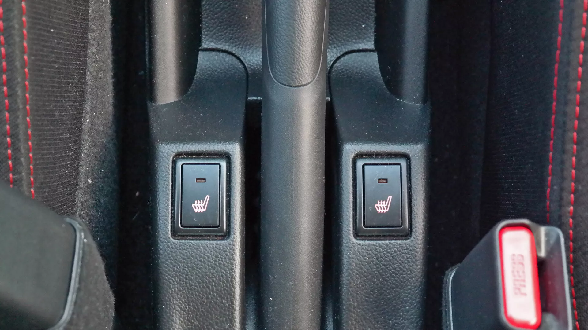 Close-up view of 2017 Suzuki Swift Sport interior center console featuring heated seat controls, textured black plastic, red stitching, and partially visible red seatbelt latch.
