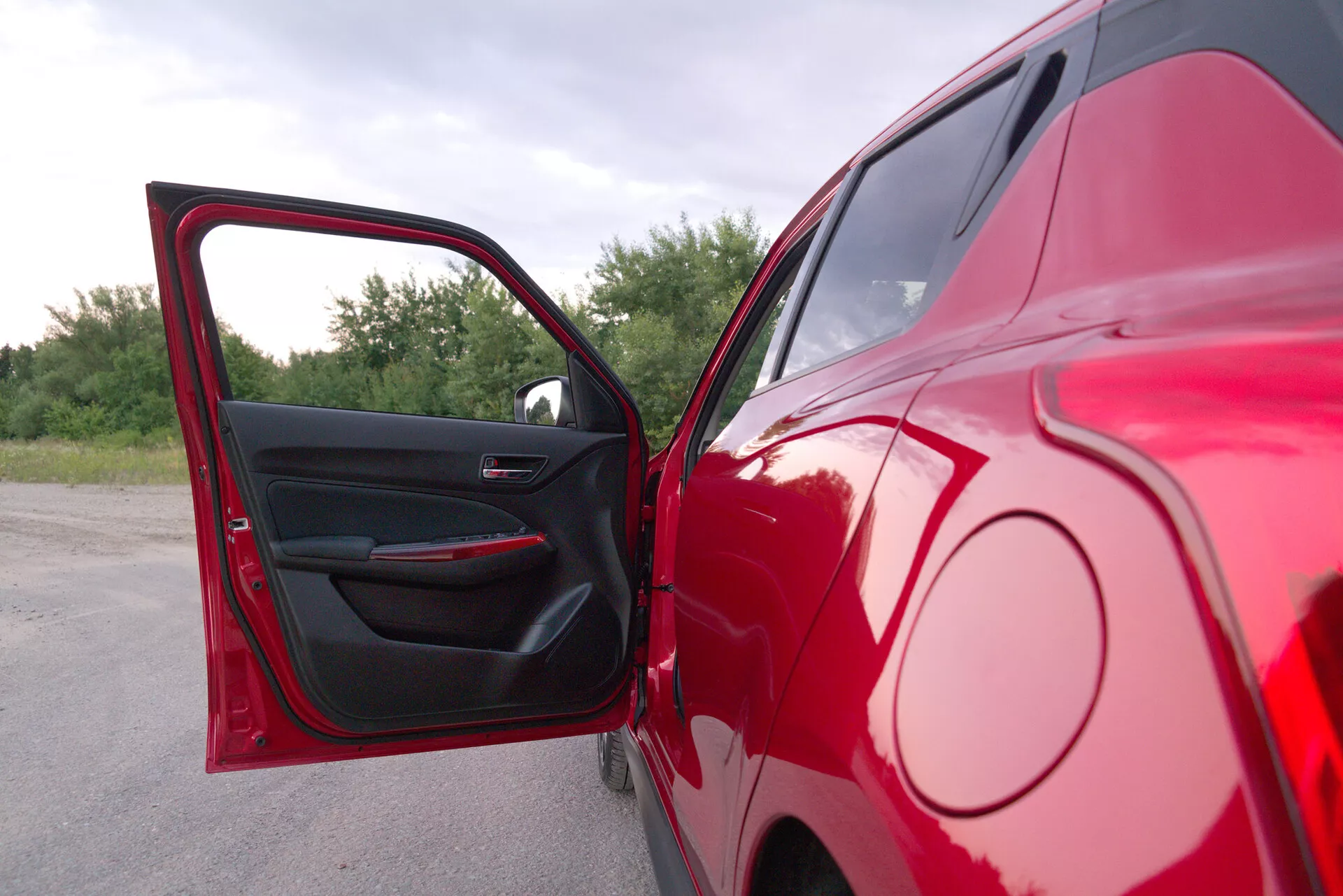 Partial side view of a 2017 Suzuki Swift Sport in red, captured from a close-to-mid-range angle with an open driver's side door. The photo highlights the rear half of the parked vehicle, showcasing a sleek glossy red exterior and a dark interior door panel with power window controls and a sleek door handle. The background features natural foliage, suggesting the car is parked outdoors.