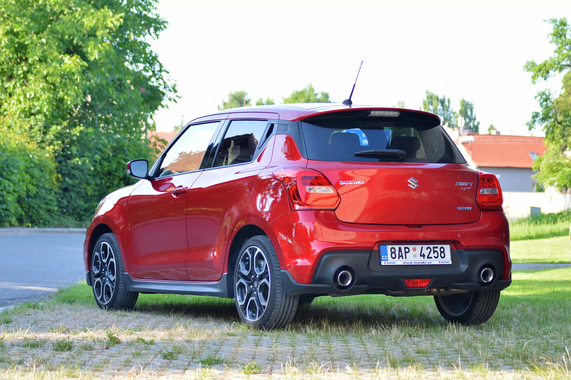 A detailed image of a modern red 2017 Suzuki Swift Sport hatchback positioned on a paved surface with grass and trees in the background. The photo is taken from a three-quarter rear perspective, showcasing the right rear side of the car. Visible features include the tail lights, rear windshield, bumper, dual-exhaust system, multi-spoke alloy wheels, and a turned rear wheel. The license plate is blurred for privacy.