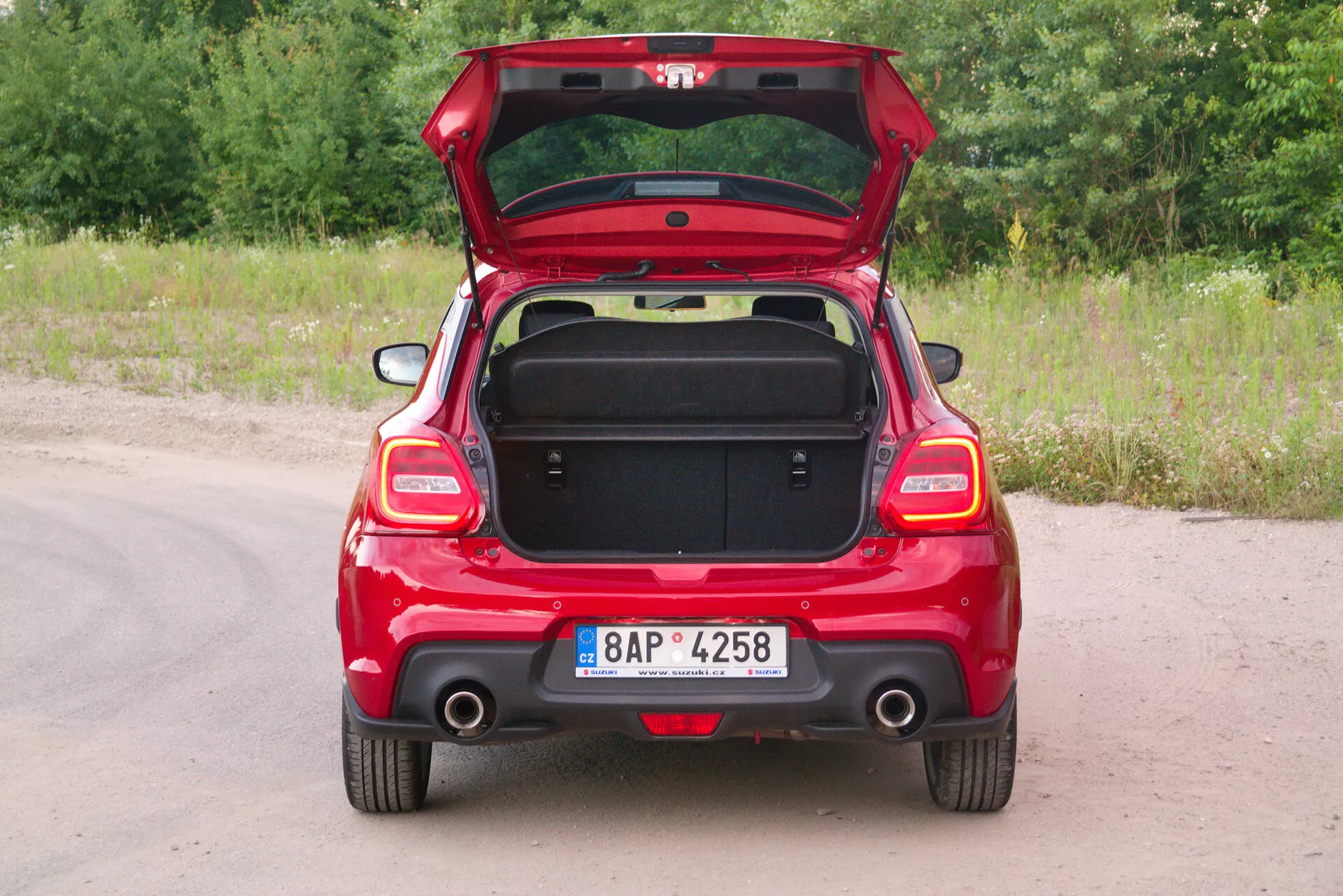 Rear view of a red 2017 Suzuki Swift Sport hatchback with the trunk lid open, showcasing symmetric back features, rear lights, single visible exhaust on passenger side, and rear windscreen wiper. The vehicle is parked on an unpaved path with grass and greenery in the background. License plate is centrally mounted on the rear bumper. The empty trunk reveals storage capacity and interior lining, with a handle grip on the inner lid for ergonomic use. Partially folded rear seats suggest a 60/40 split for expandable cargo space. Simple rear bumper indicates standard features without advanced technology add-ons.