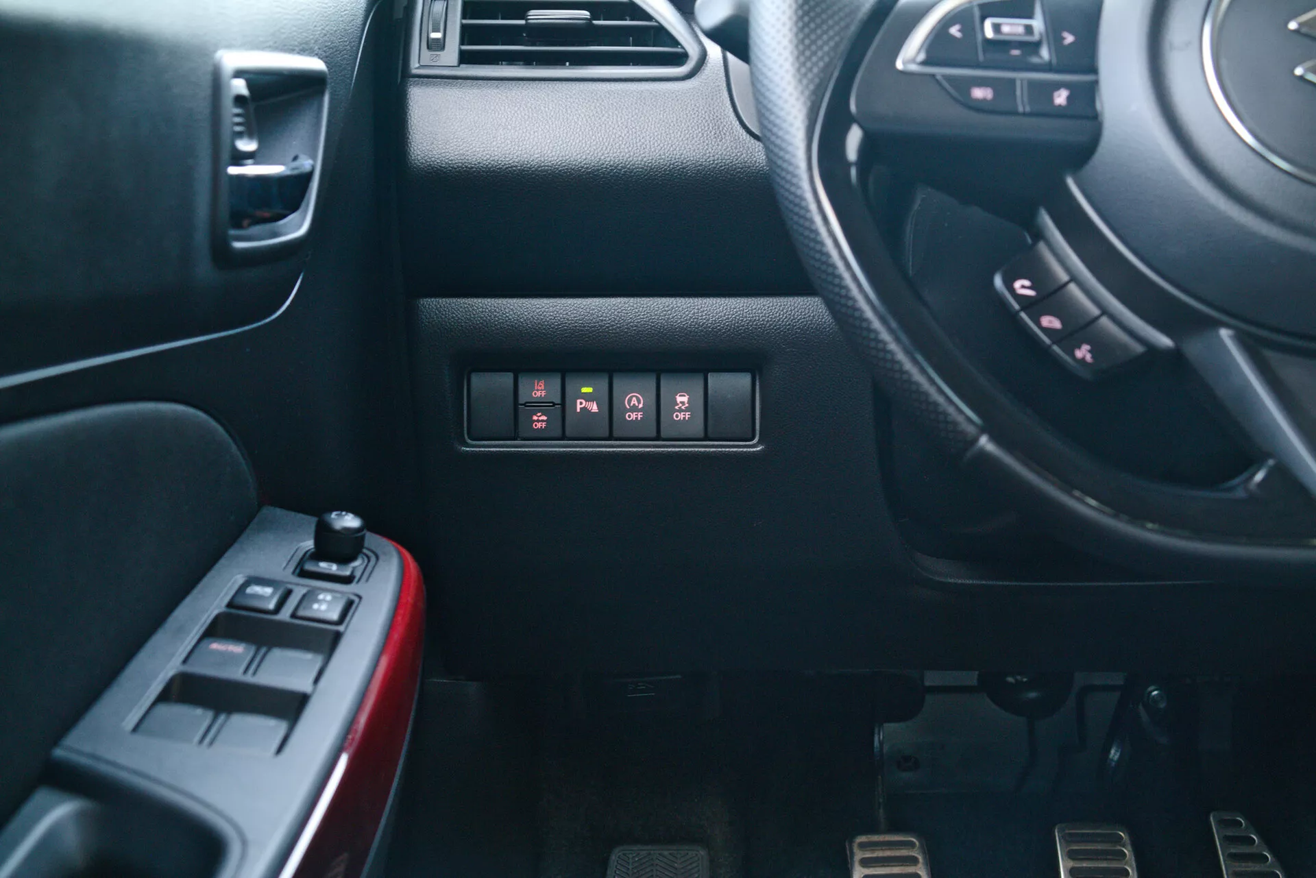 Interior view of a 2017 Suzuki Swift Sport from the passenger's perspective, showcasing the driver's dashboard and side of the center console. View includes partial steering wheel with multifunction buttons, dashboard with push-button switches, driver's door with power window and mirror controls, sporty metal pedals for a manual transmission, and detailed textures and materials.