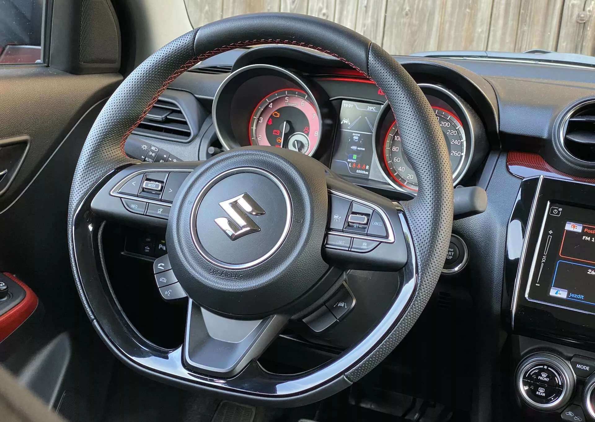 Interior view of a 2017 Suzuki Swift Sport from the driver's seat perspective, showcasing the sporty three-spoke, flat-bottom steering wheel with multifunctional buttons for multimedia and convenience technology, including volume adjustment, voice command, menu navigation, and cruise control. The steering wheel is wrapped in leather with contrasting stitch details and features the Suzuki emblem prominently. Behind it, the instrument cluster displays a central analog speedometer, tachometer, fuel gauge, and a small digital display for additional vehicle data. The dashboard integrates air vents and an infotainment touchscreen with audio and air conditioning controls. The color scheme is predominantly black with metallic trim and red accents, underscoring a sporty aesthetic.
