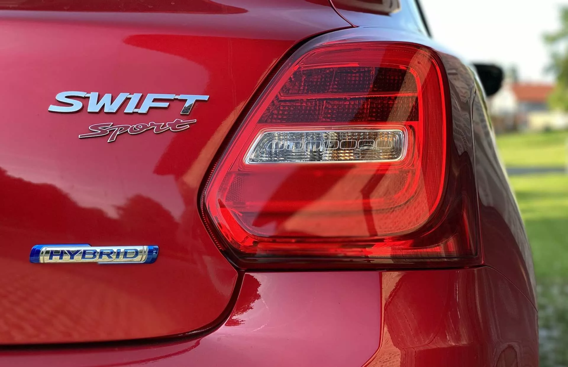 Close-up view of the rear portion of a red Suzuki Swift Sport 2017, focusing on the driver's side LED tail light assembly. The image captures the glossy finish of the car's paint, the SWIFT Sport and HYBRID badges, and the contours of the rear fender and part of the trunk lid. The shallow focus blurs the background and highlights the details of the light assembly and badges, emphasizing the car's modern design and hybrid technology.