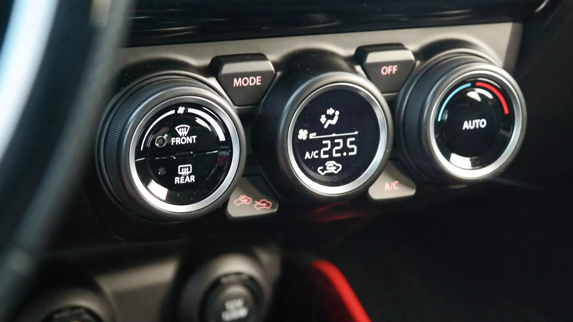 Close-up view of the 2017 Suzuki Swift Sport climate control system, taken from the driver's seat perspective. The image highlights the center console with three black circular knobs featuring white and red accents; the left knob labeled 'MODE' controls airflow direction, the middle knob adjusts the power and fan speed with an 'OFF' position, and the right knob shows a digital temperature readout of '22.5' degrees Celsius indicating automatic climate control with 'AUTO' and 'AC' functions. Below these, additional buttons are partially visible, including one with a defrost symbol. The background is blurred to emphasize the climate controls.