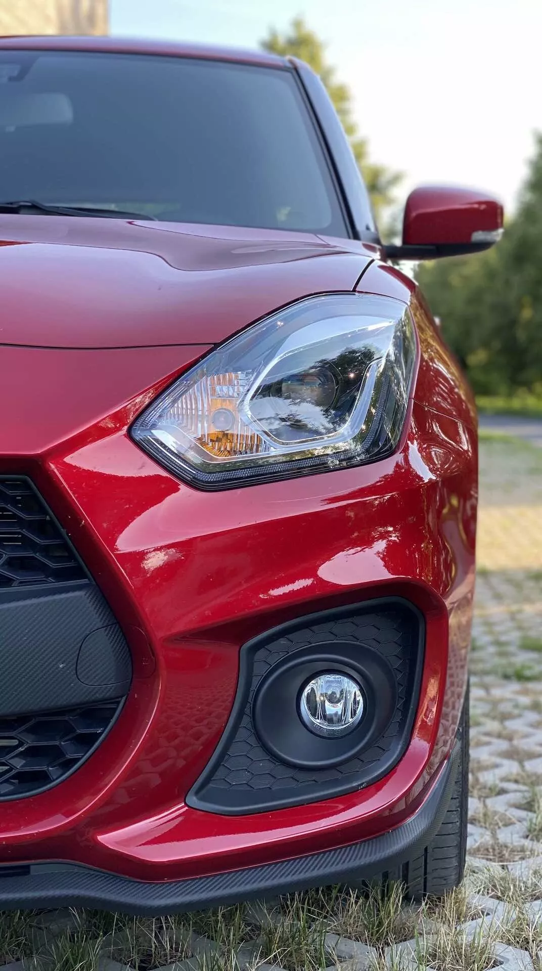 Close-up view of a red 2017 Suzuki Swift Sport from a low angle, focusing on the front-left portion featuring the driver's side headlight with intricate designs potentially inclusive of LED or halogen lighting technology, partially blurred emblem on the grille, and streamlined side mirror set against a natural background with greenery.