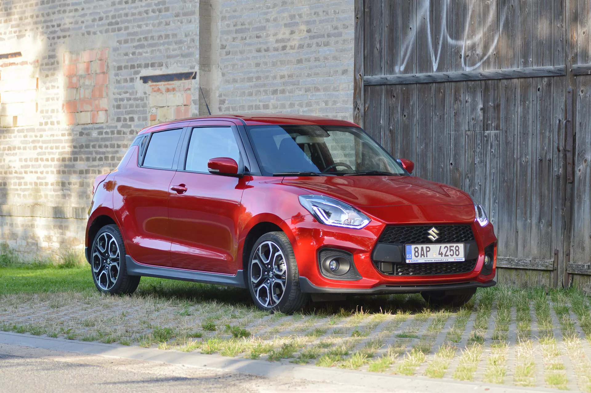 Image of a red Suzuki Swift Sport 2017 hatchback captured from a three-quarter front angle, highlighting the front and side profiles. The car's design features include a prominent front grille with integrated logo, sleek headlights, fog light housings, and multi-spoke alloy wheels. The shot is taken from a slight height, providing a top-down view of the hood and roof. The surroundings include a grassy foreground and a brick wall with graffiti in the background, emphasizing the sporty and dynamic appearance of the vehicle.
