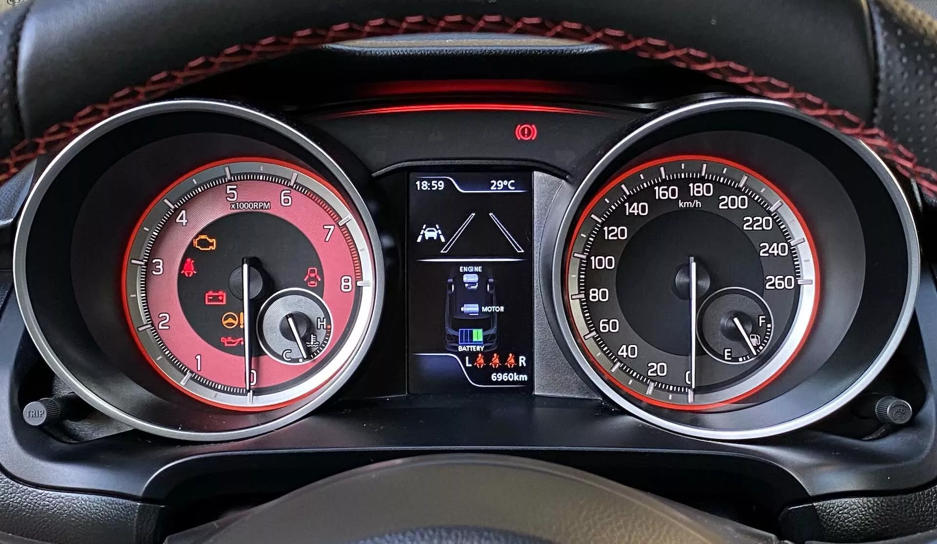 Close-up view of the instrument cluster in a 2017 Suzuki Swift Sport from the driver's seat perspective. The image showcases the dual-gauge layout with a red tachometer (up to 8,000 RPM) on the left and a matching red-themed speedometer (up to 260 km/h) on the right, with chrome accents and white numbering for enhanced visibility. A central small digital display shows the time (18:59), exterior temperature (29°C), reverse gear selection, and odometer reading (96604 km). Additional warning lights are located on the top border, alongside red stitching that adds a sporty touch.