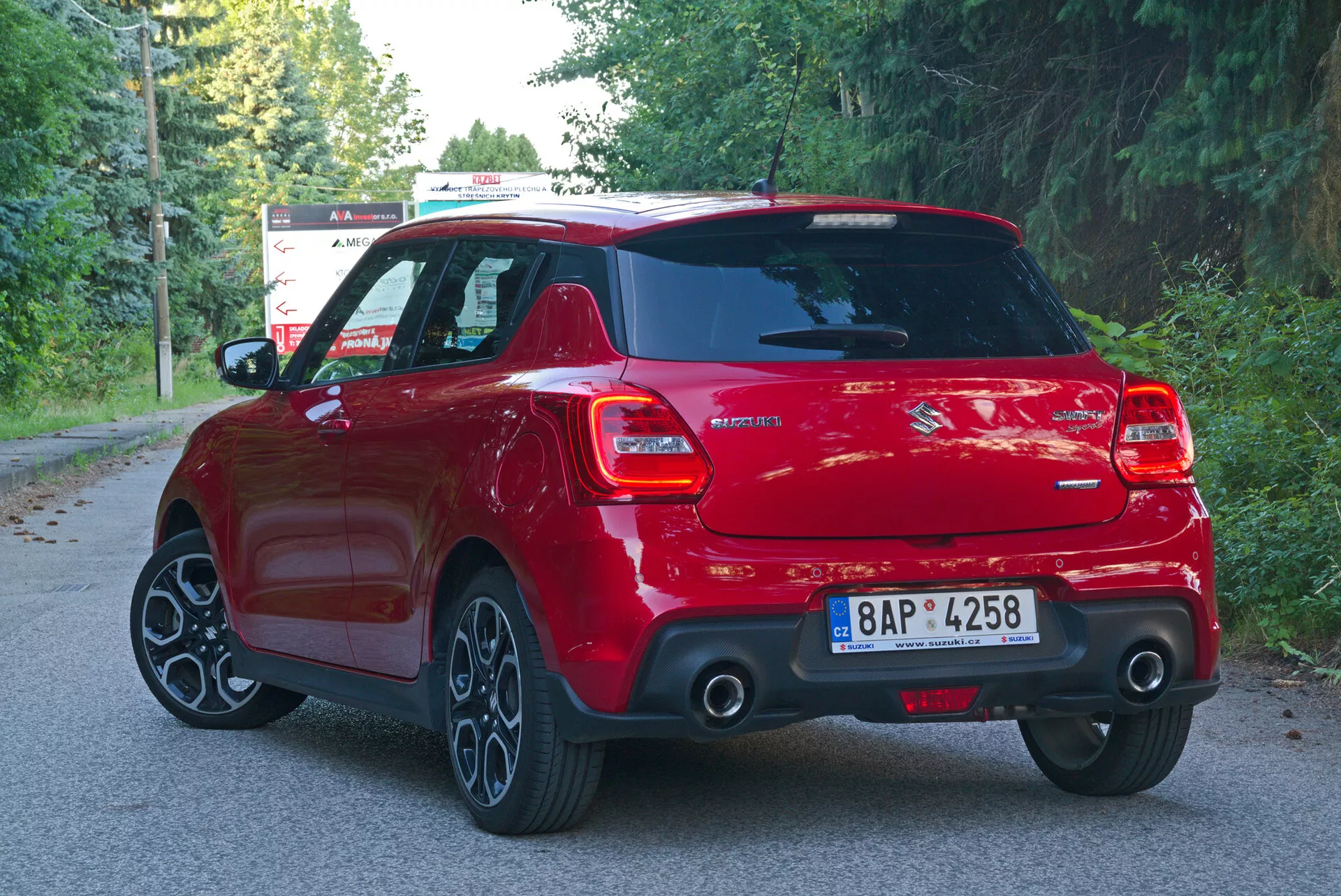 This image shows a rear three-quarter angle view of a red Suzuki Swift 2017 (Swift-Sport) hatchback car, capturing the back right side of the vehicle. The shot highlights the car's modern sporty design with distinctive features such as LED tail lights, a rear windshield wiper, a central rear badge with model information, a visible exhaust pipe, a roof spoiler, multi-spoke alloy wheels, and a rear bumper equipped with a parking sensor system. The well-lit photo taken in natural daylight suggests the vehicle is parked on a suburban road lined with trees, with its roof and antenna clearly visible. The license plate has been blurred for privacy.